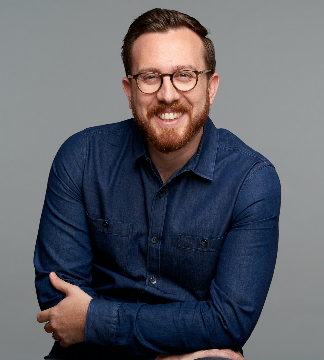 Portrait of our advisor, Ben Ramirez, a man with glasses, red hair, and a beard smiling, wearing a blue button-up shirt, against a gray background.