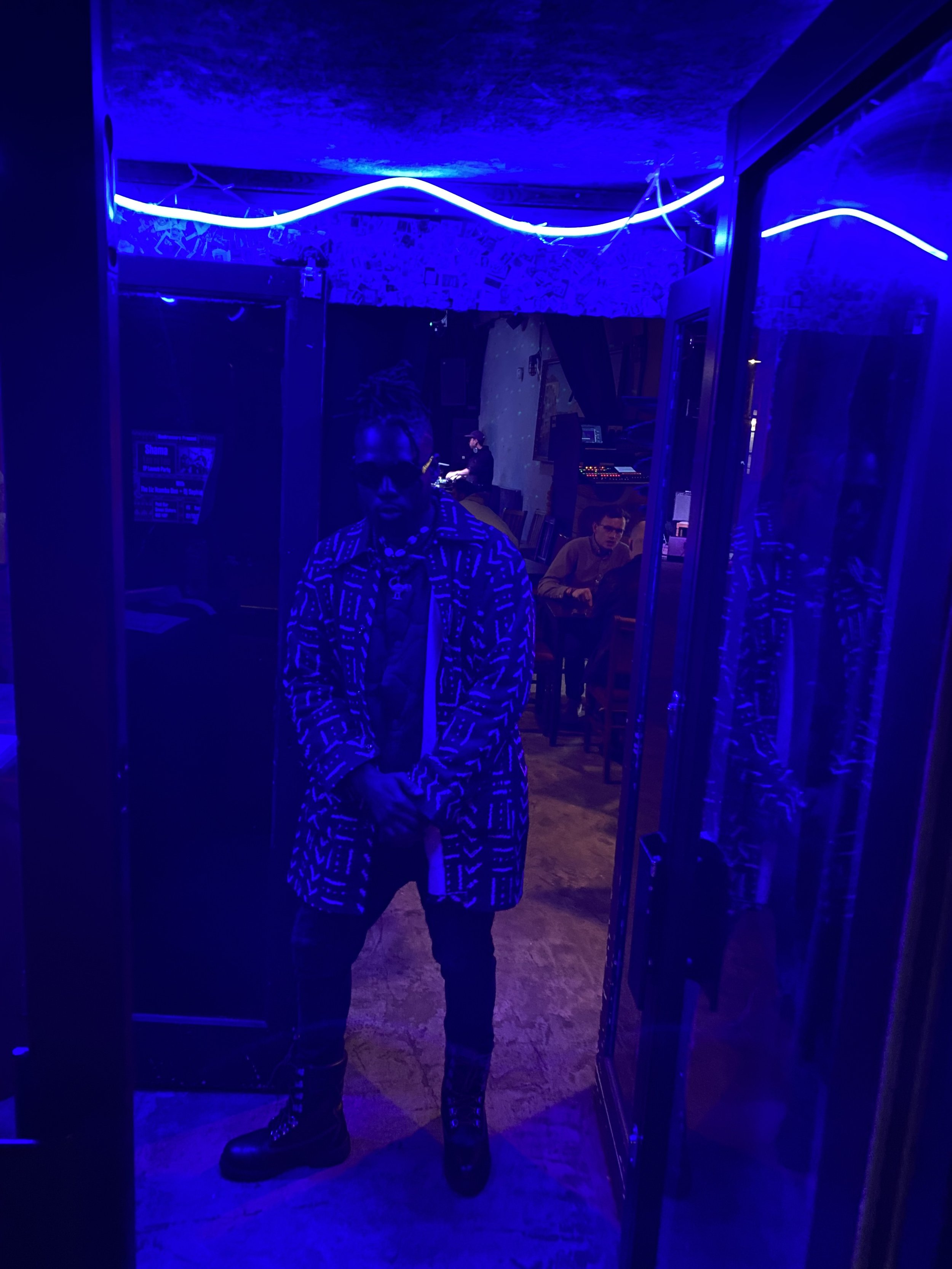 Man in patterned coat and boots standing under blue lighting in a dimly lit room with neon lights.