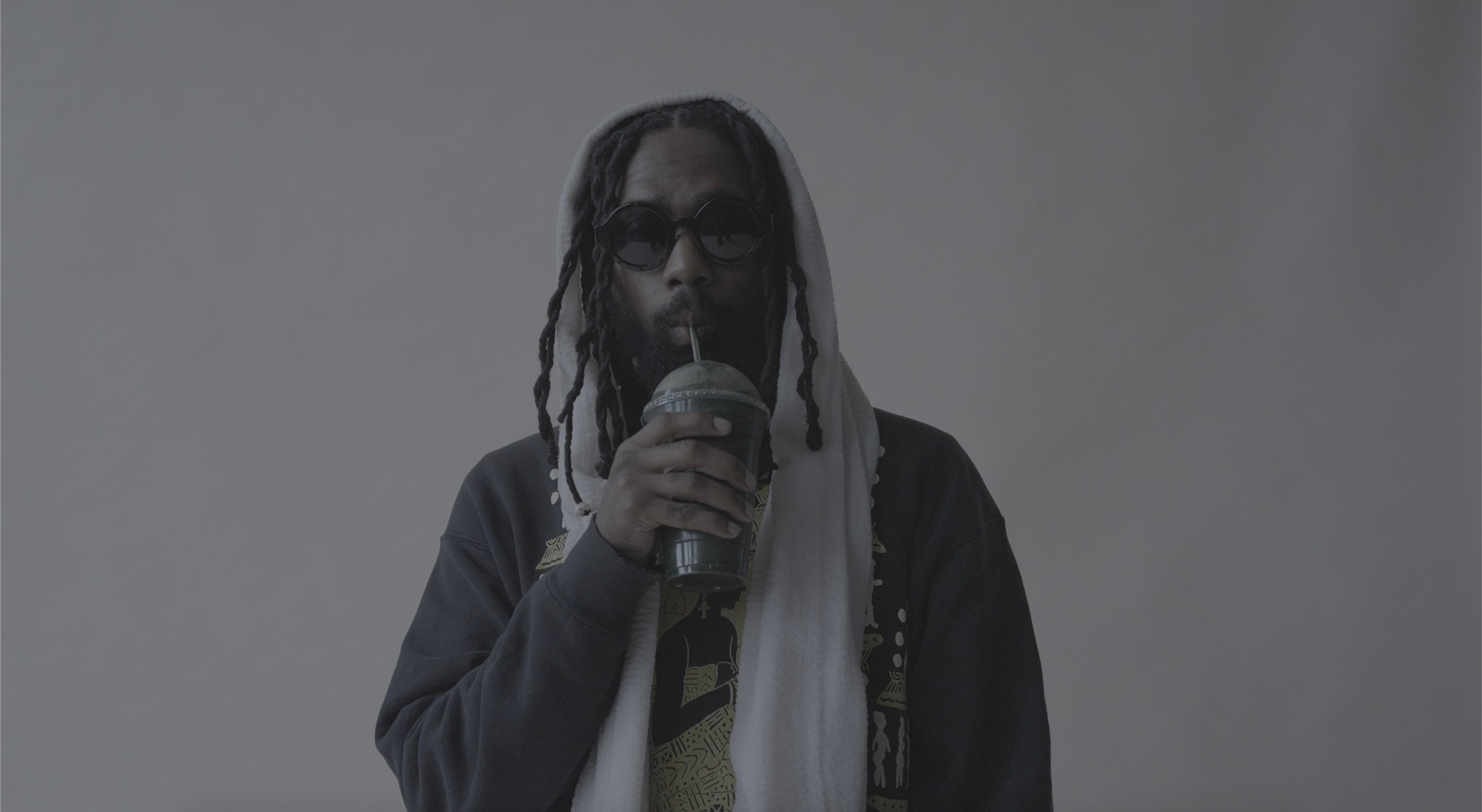 Person with dreadlocks wearing sunglasses and a hood, holding a smoothie with a straw against a plain background.