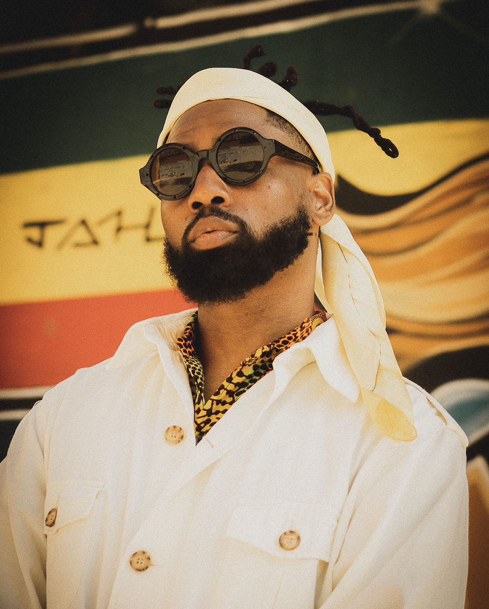 A man wearing round sunglasses, a headscarf, and a white button-up shirt, standing in front of a colorful mural.