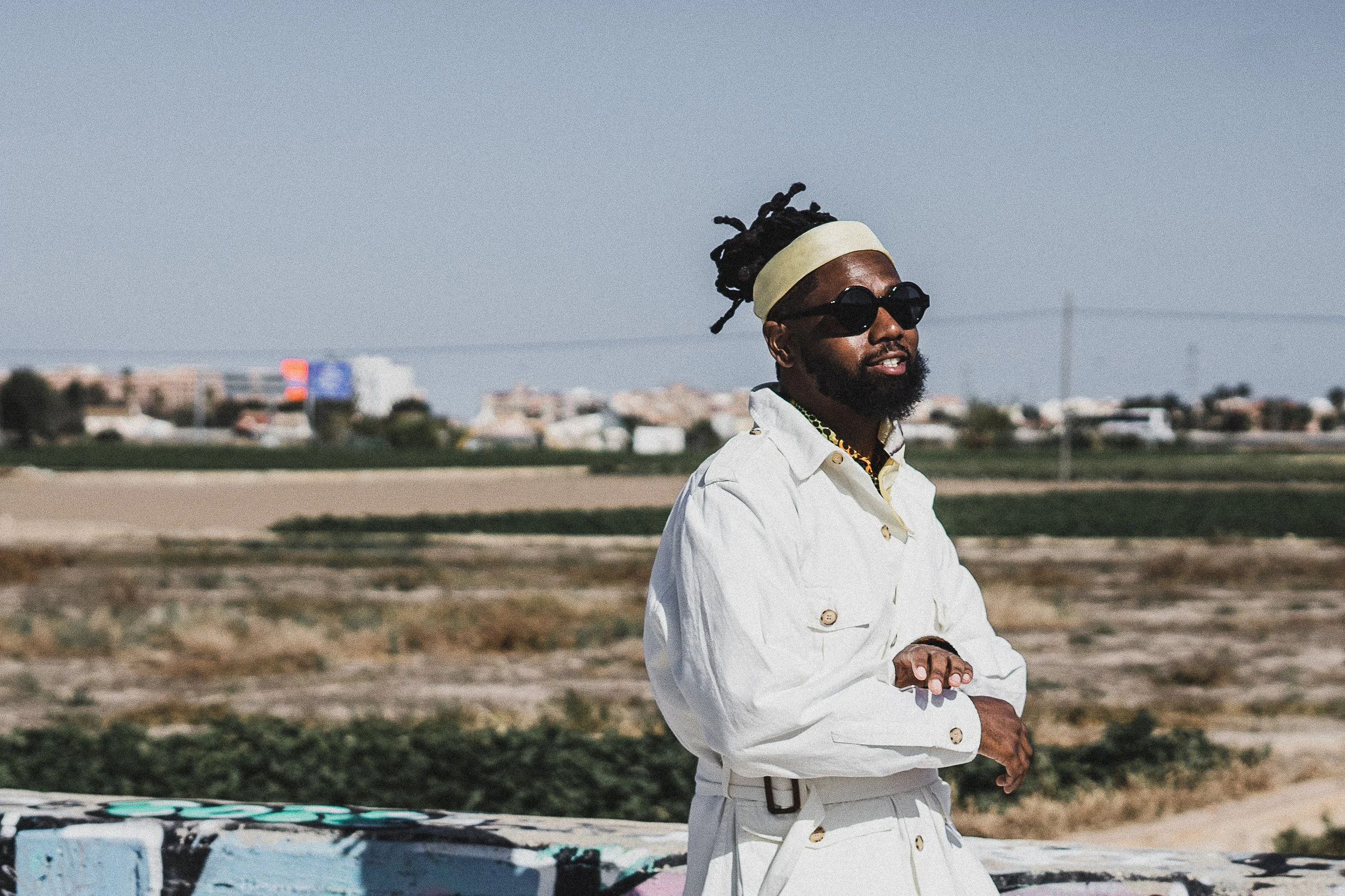 Person in white trench coat and sunglasses standing outside, with graffiti-covered barrier and blurred landscape in the background.