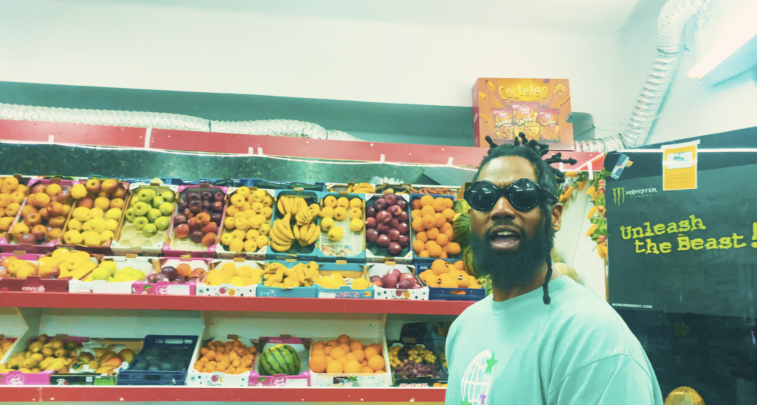 Man wearing sunglasses in front of a fruit display at a store, featuring bananas, oranges, apples, and lemons.
