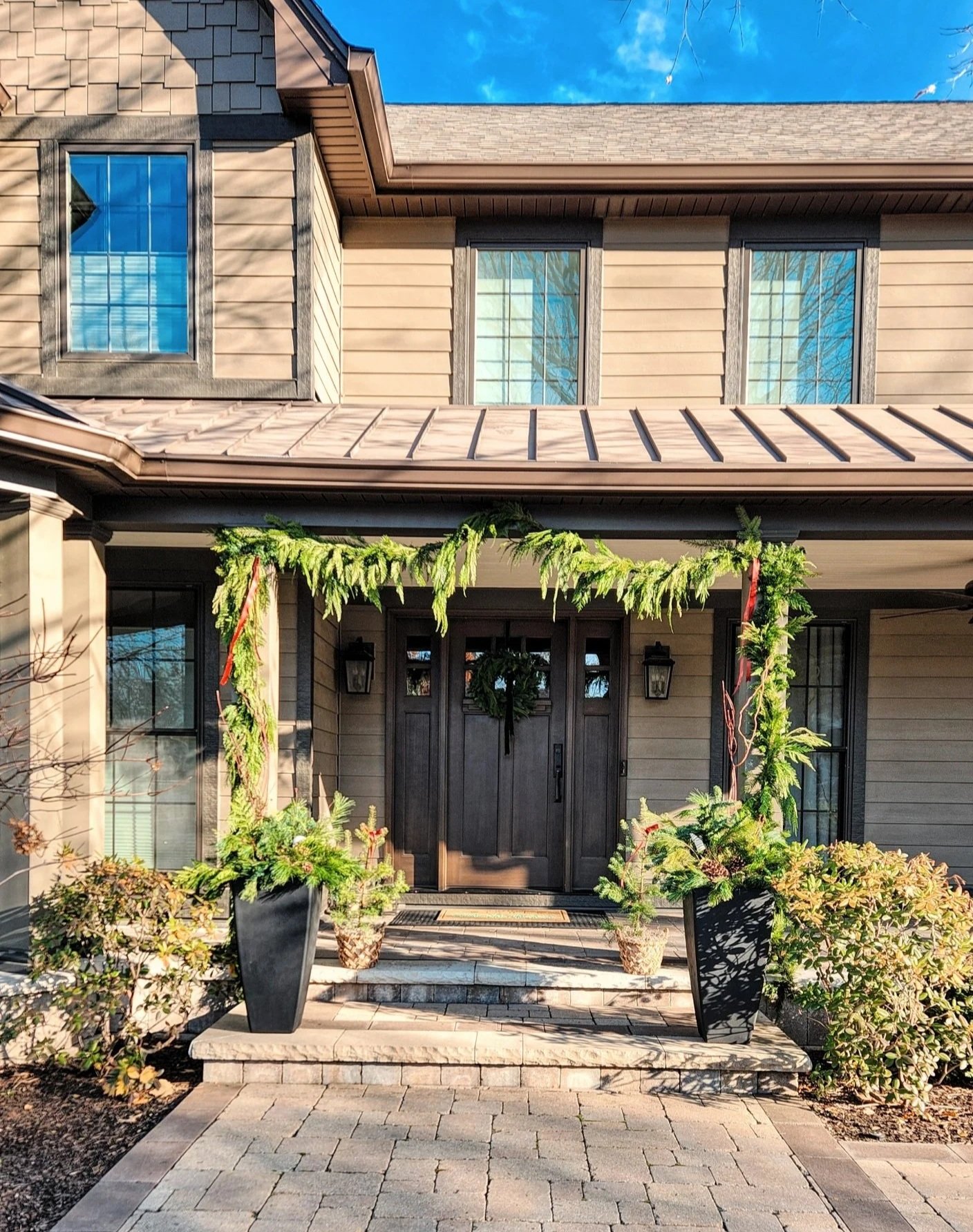Decorated house entrance with a black door, wreath, potted plants, and greenery arch, under a blue sky.