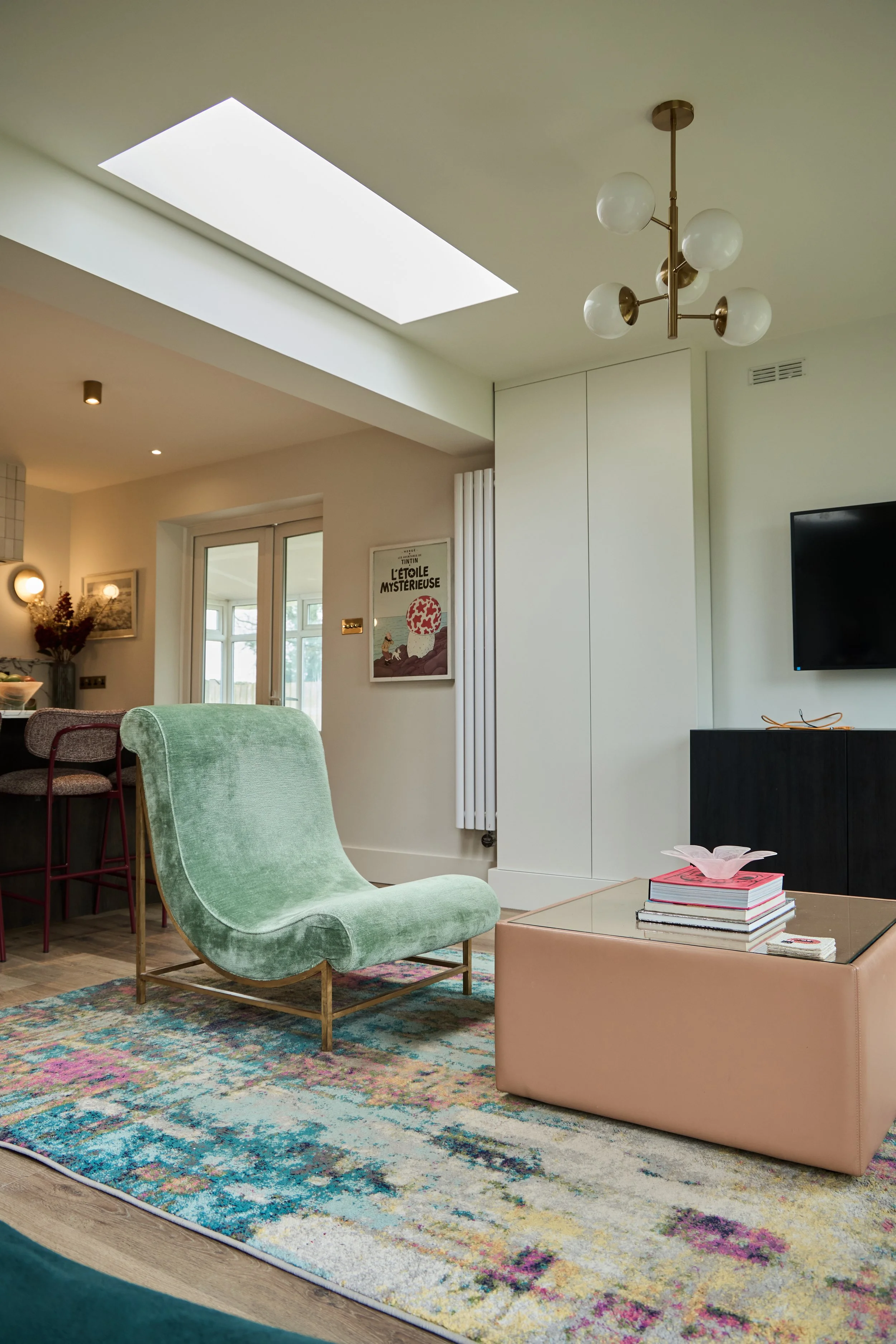 Modern living room with a skylight, a green lounge chair, a pink ottoman with books and a decorative bowl, a colorful area rug, and a wall-mounted television.