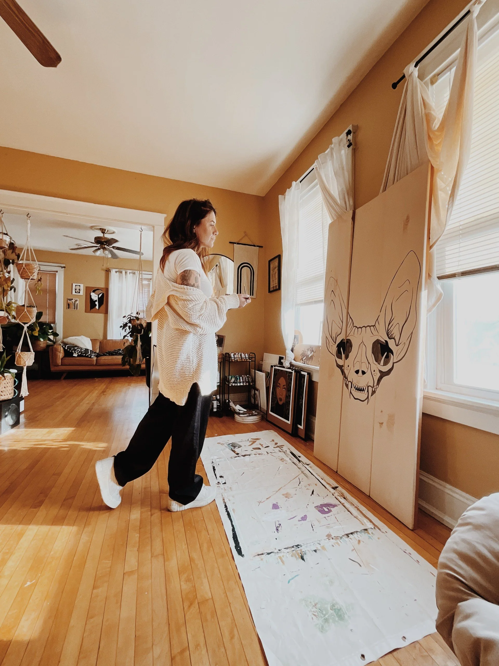 A woman standing in a cozy living room, painting a large canvas with a skull and horns design, natural light coming through windows, hardwood floors, art and furniture in the background.