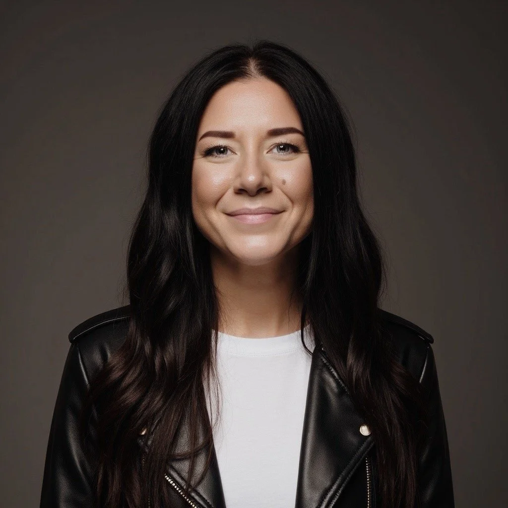 Portrait of a woman with long dark hair, smiling, wearing a black leather jacket and a white top, against a gray background.