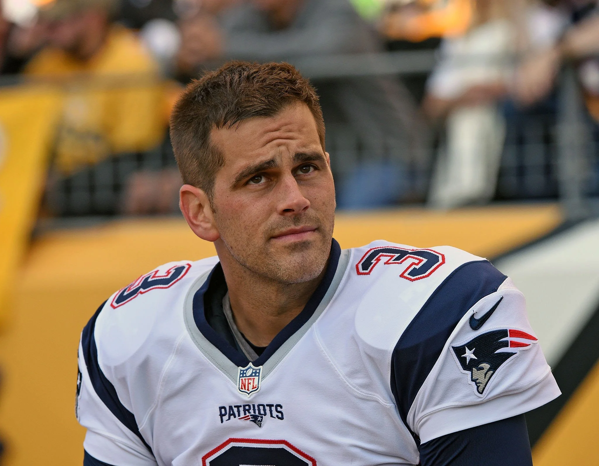 A football player in a New England Patriots uniform sitting on the sideline with a thoughtful expression.