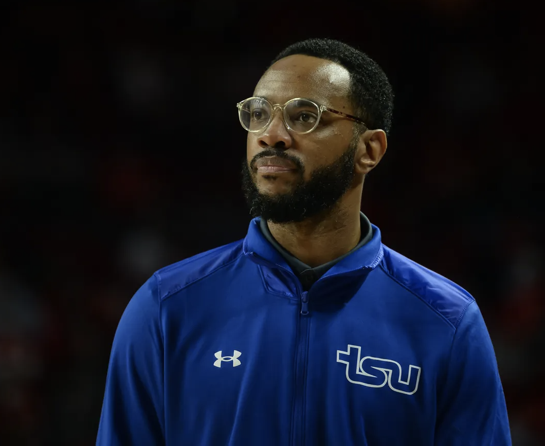 A man with glasses and a beard wearing a blue LSU jacket, standing indoors.