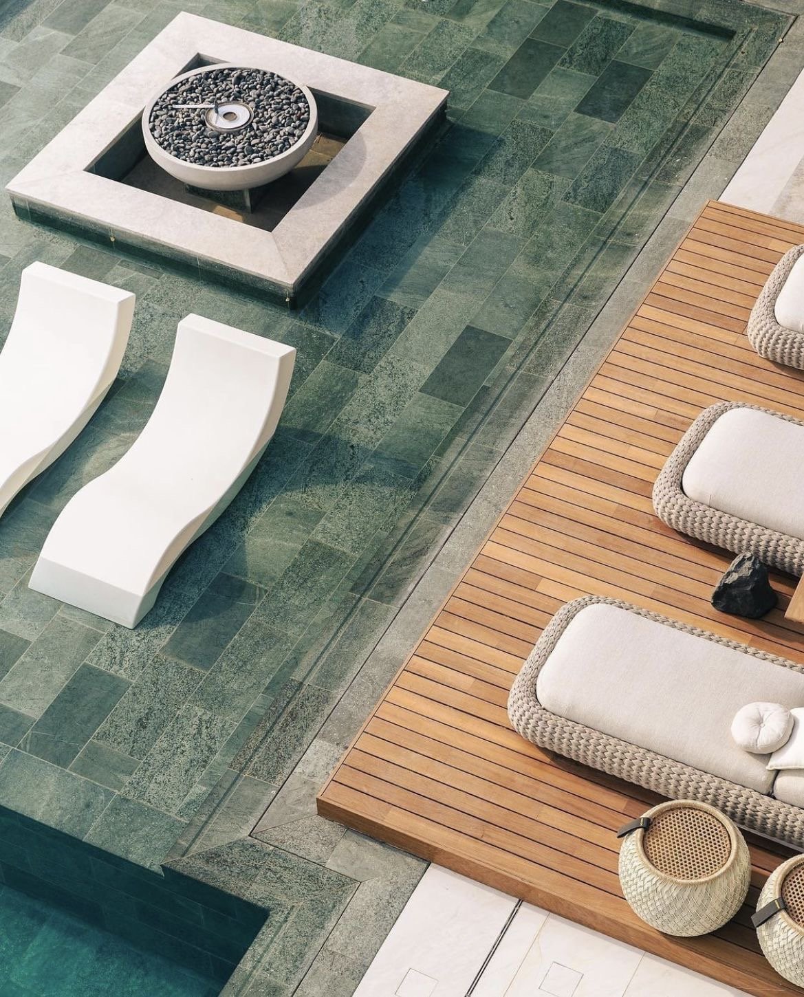 A poolside area with two white modern lounge chairs, a wooden deck with beige cushioned seating, a small round side table, and a rectangular water fountain with a stone base, all surrounded by green stone tiles.