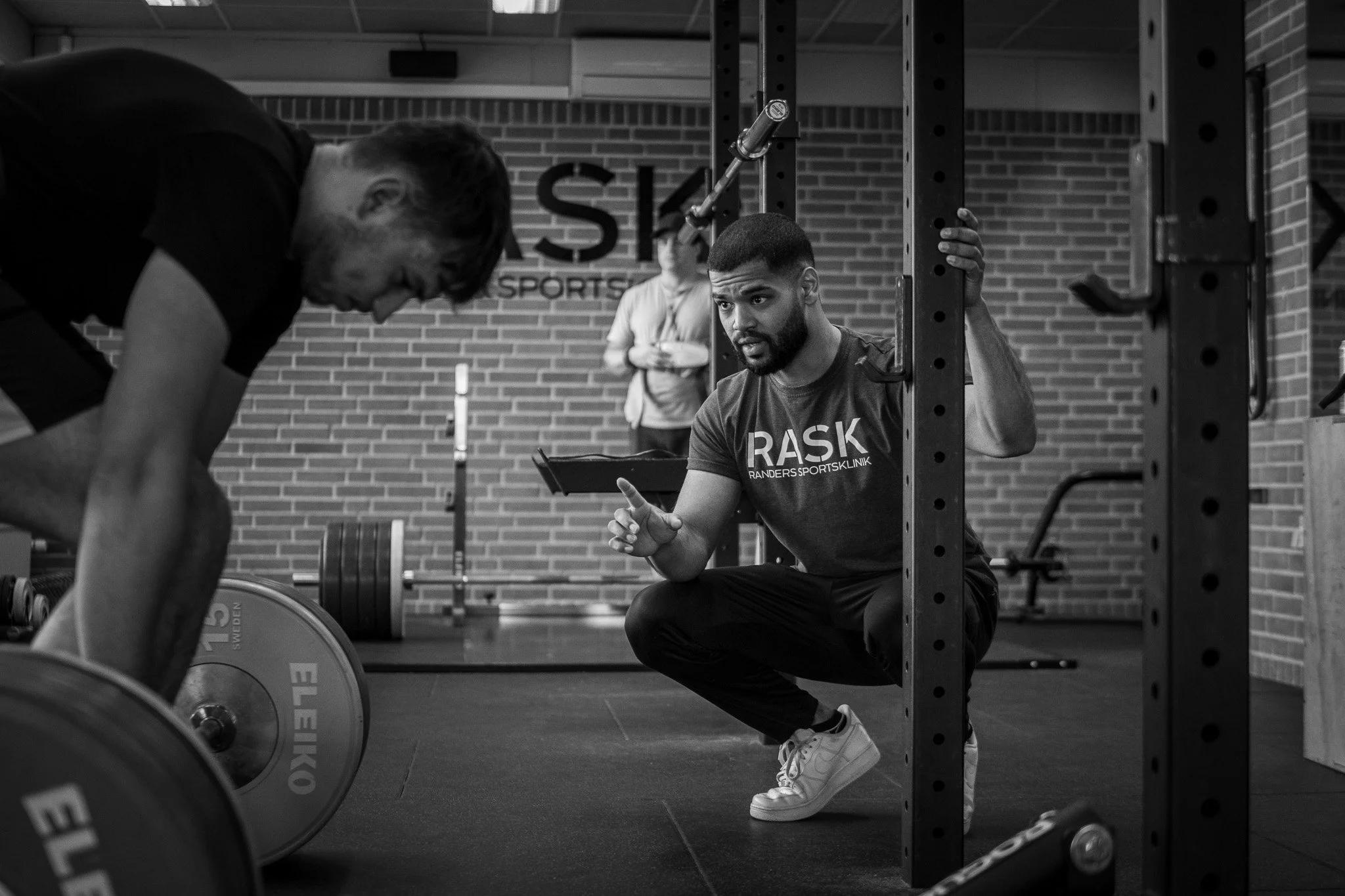 A personal trainer kneeling and explaining exercise form to a man lifting weights in a gym.