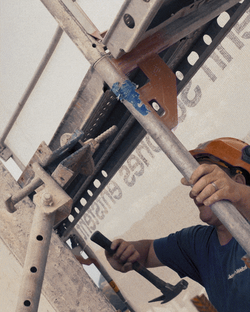 Worker using a hammer and holding a mobile device on a construction scaffold.