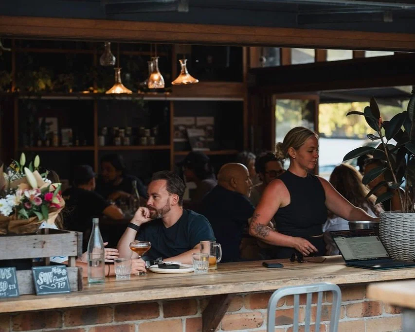 Interior of a lively Roadhouse Byron Bay lunch with patrons dining and a waitress serving at a bar, with warm lighting and a brick counter.
