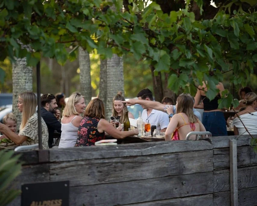 Group of people enjoying outdoor dining and happy hour at Roadhouse Byron Bay under trees at a social gathering with food and drinks on the table.
