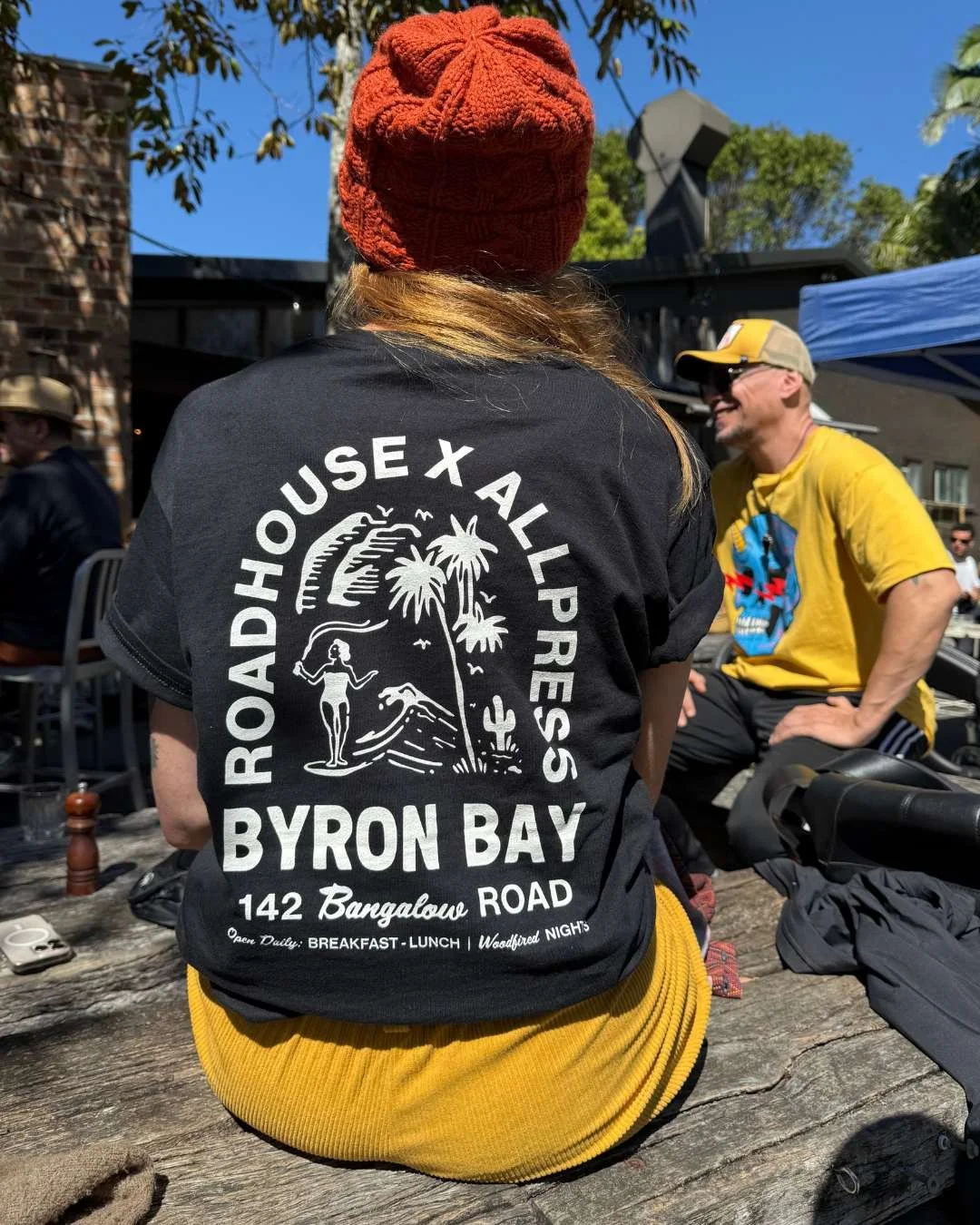 Two people sitting at The Roadhouse Byron Bay in the sun wearing Perk Up Byron tee shirts