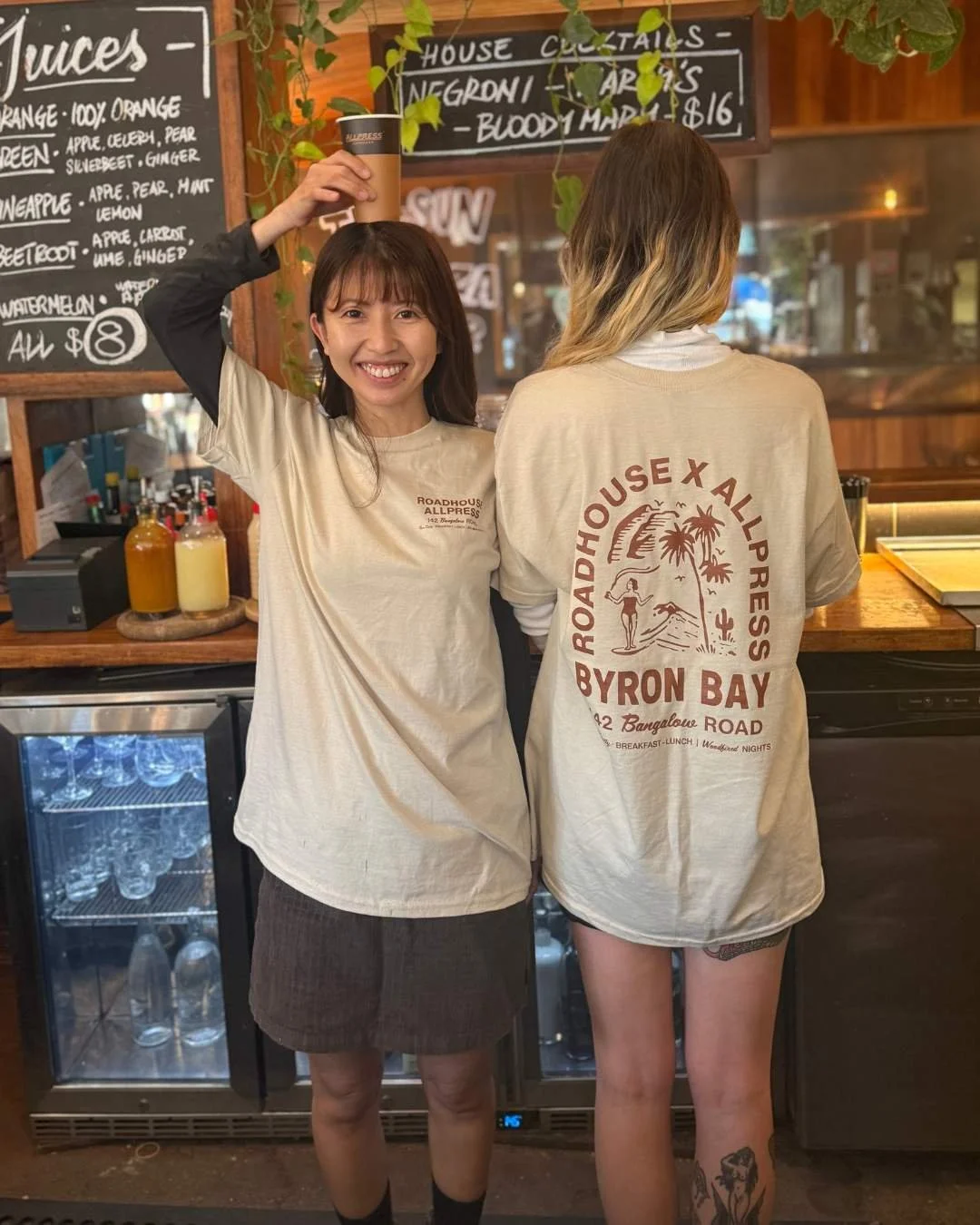 Two Roadhouse staff wearing event tees and holding a coffee cup on their heads