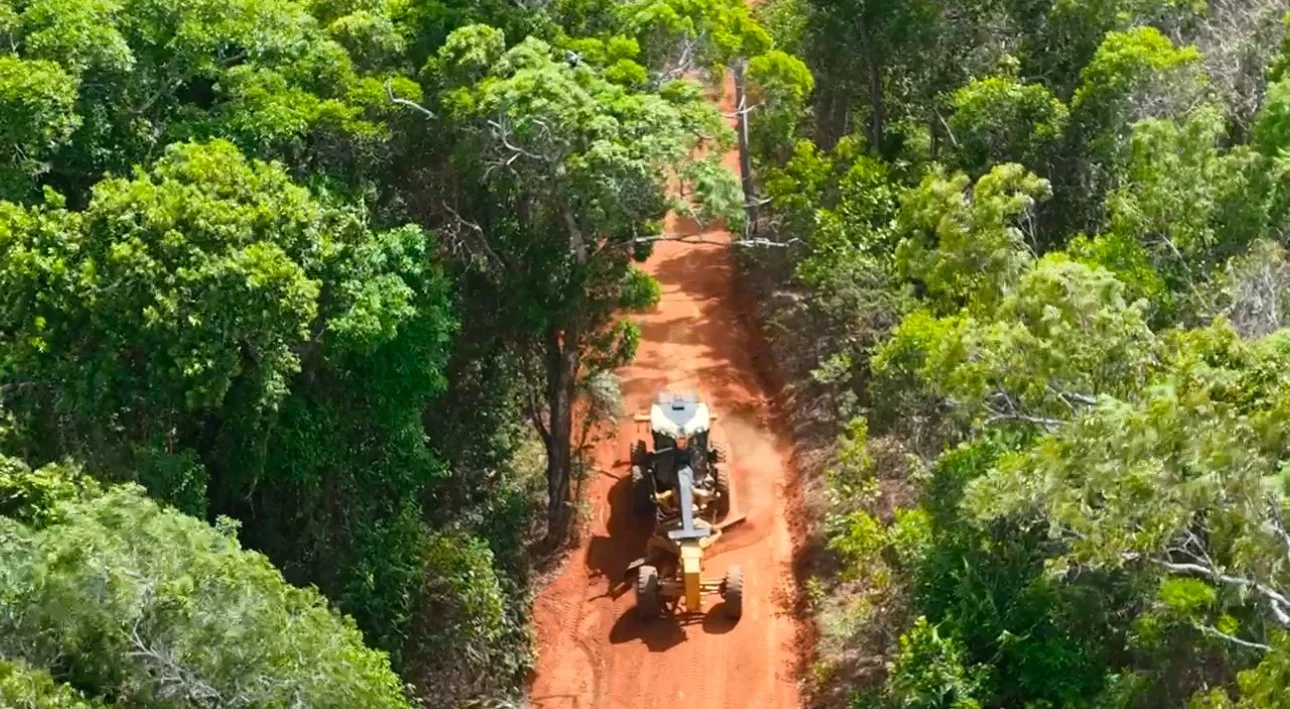 An aerial view of a tractor working on a red dirt road through a dense green forest.