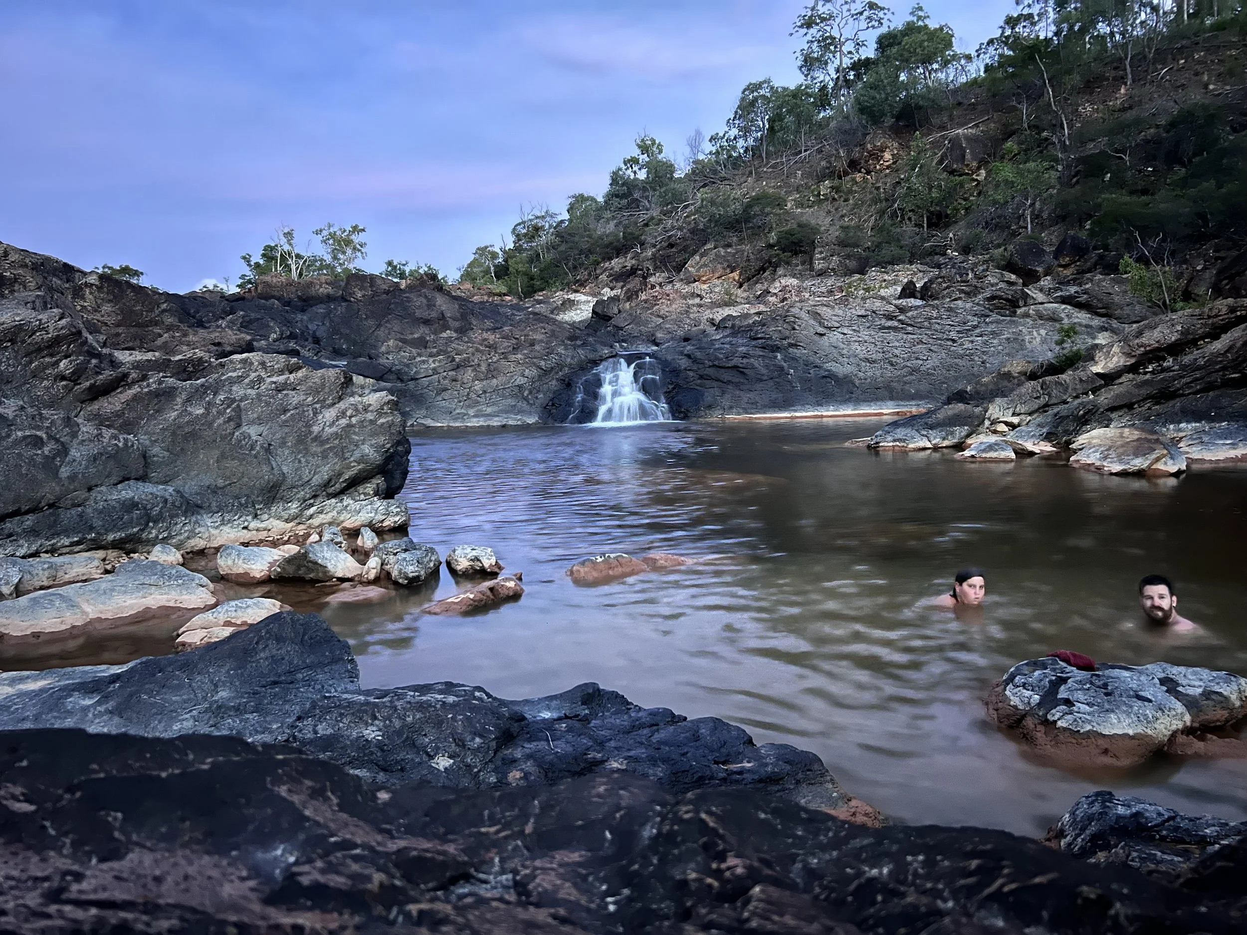 Two people swimming in a natural rock pool with a small waterfall and rocky cliffs in the background.