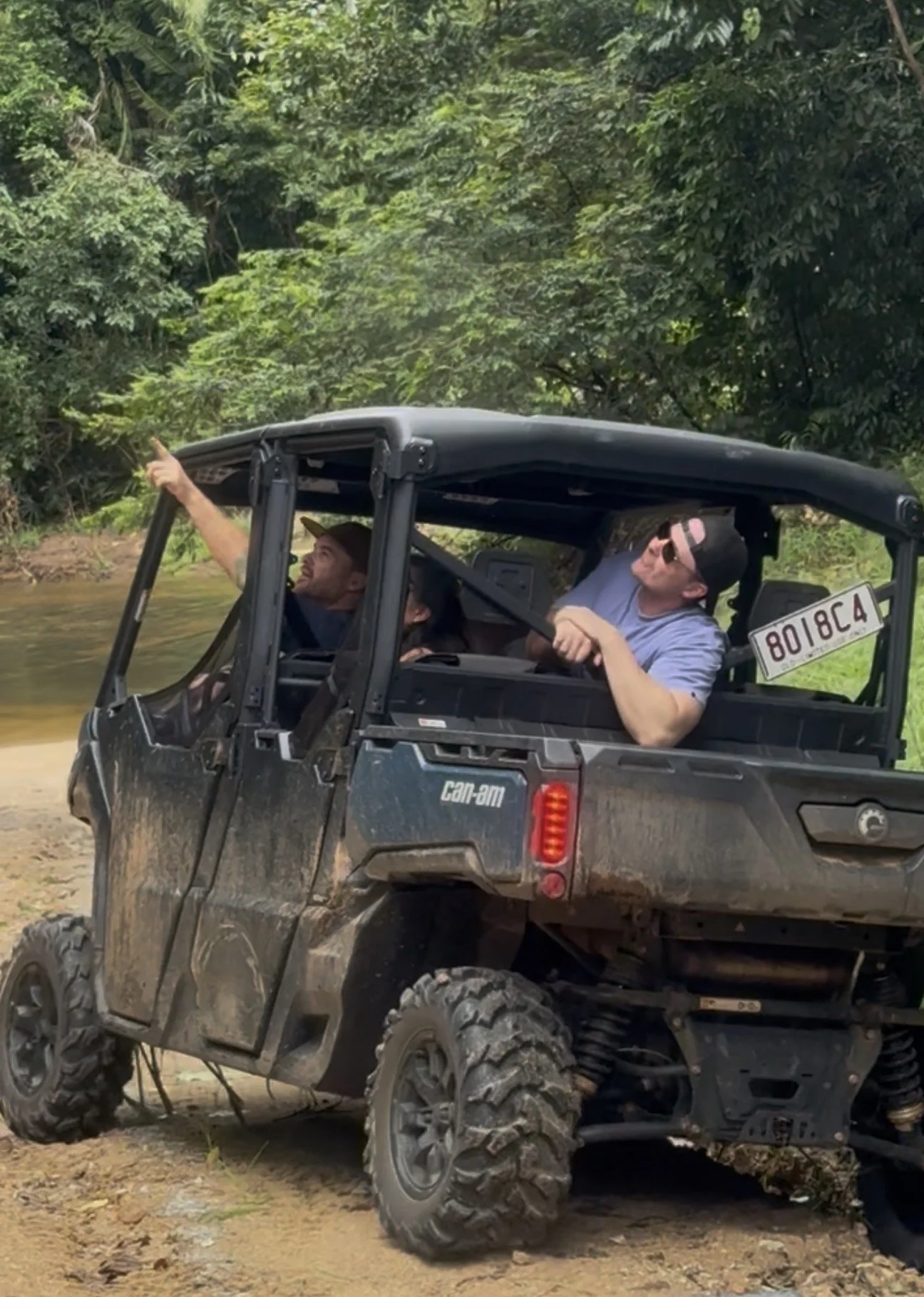 Two men riding in a side-by-side utility vehicle in a forested area near a body of water, with one pointing and the other wearing sunglasses.