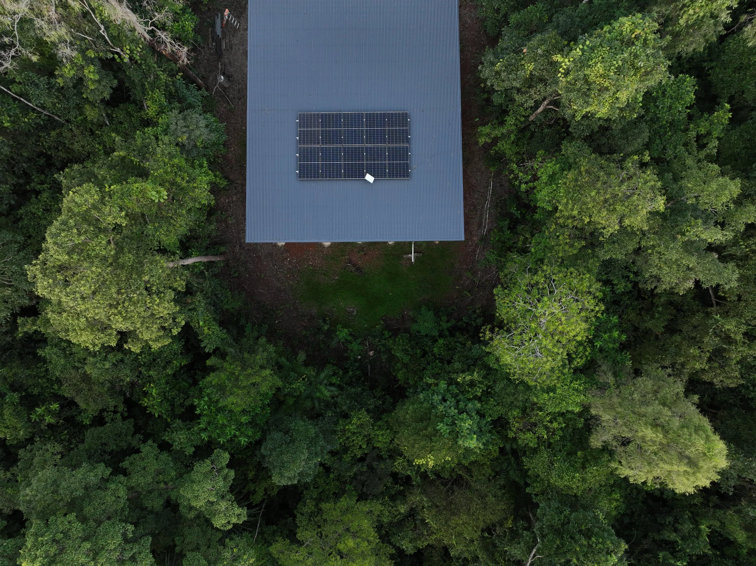 Aerial view of a house's roof fitted with solar panels, surrounded by a dense green rainforest.