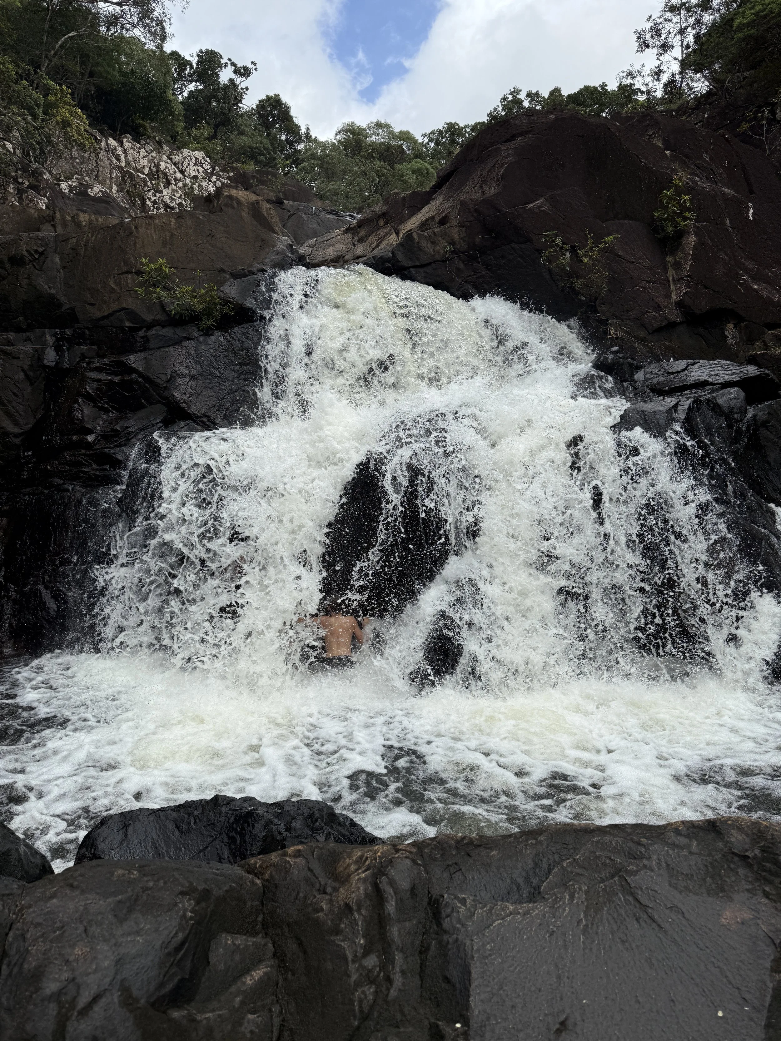 Person swimming under a waterfall surrounded by rocks and trees.