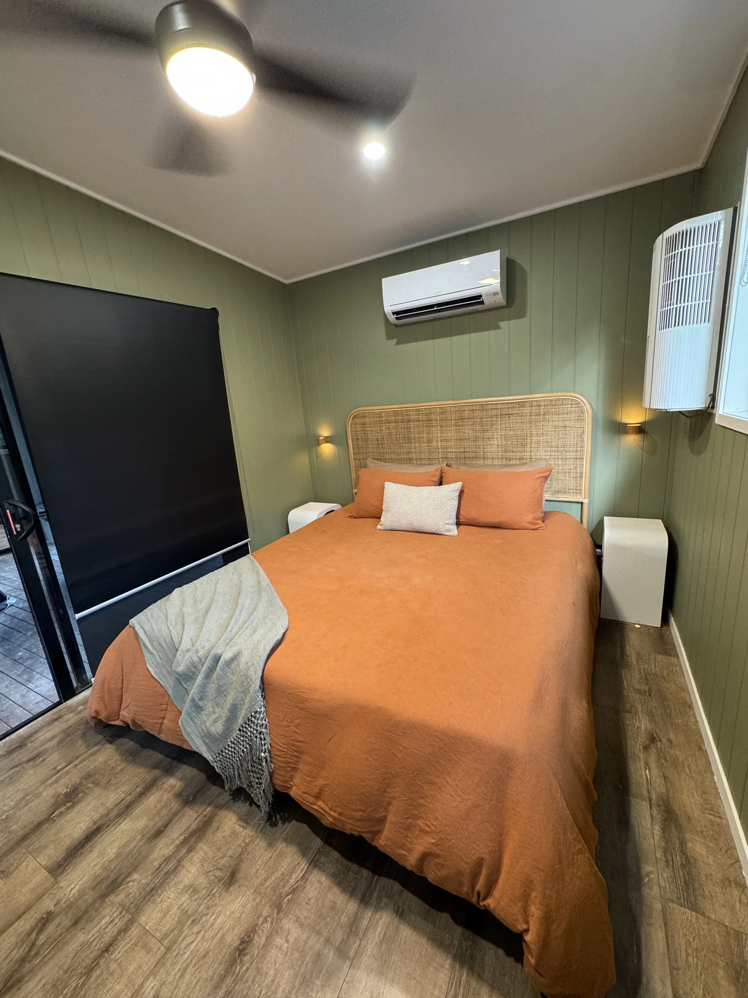 A cozy bedroom with a green paneled wall, a bed with orange bedding and pillows, a woven headboard, two bedside tables, a wall-mounted air conditioner, a window with open blinds, a ceiling fan, and hardwood flooring.