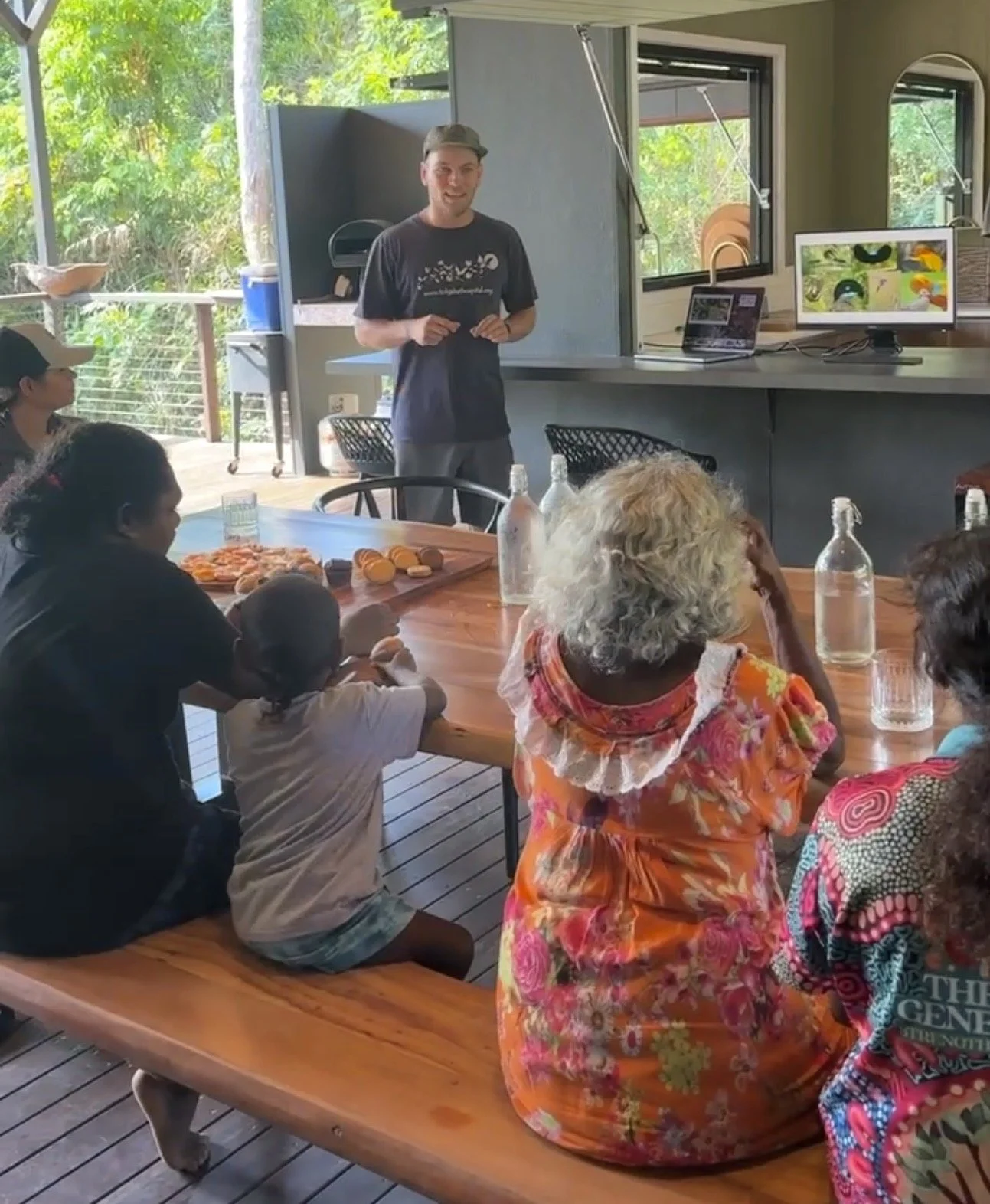 A group of children and adults sitting around a wooden table, listening to a young man speaking in a bright, open room with large windows showing greenery outside. There are bottles of water and snacks on the table.