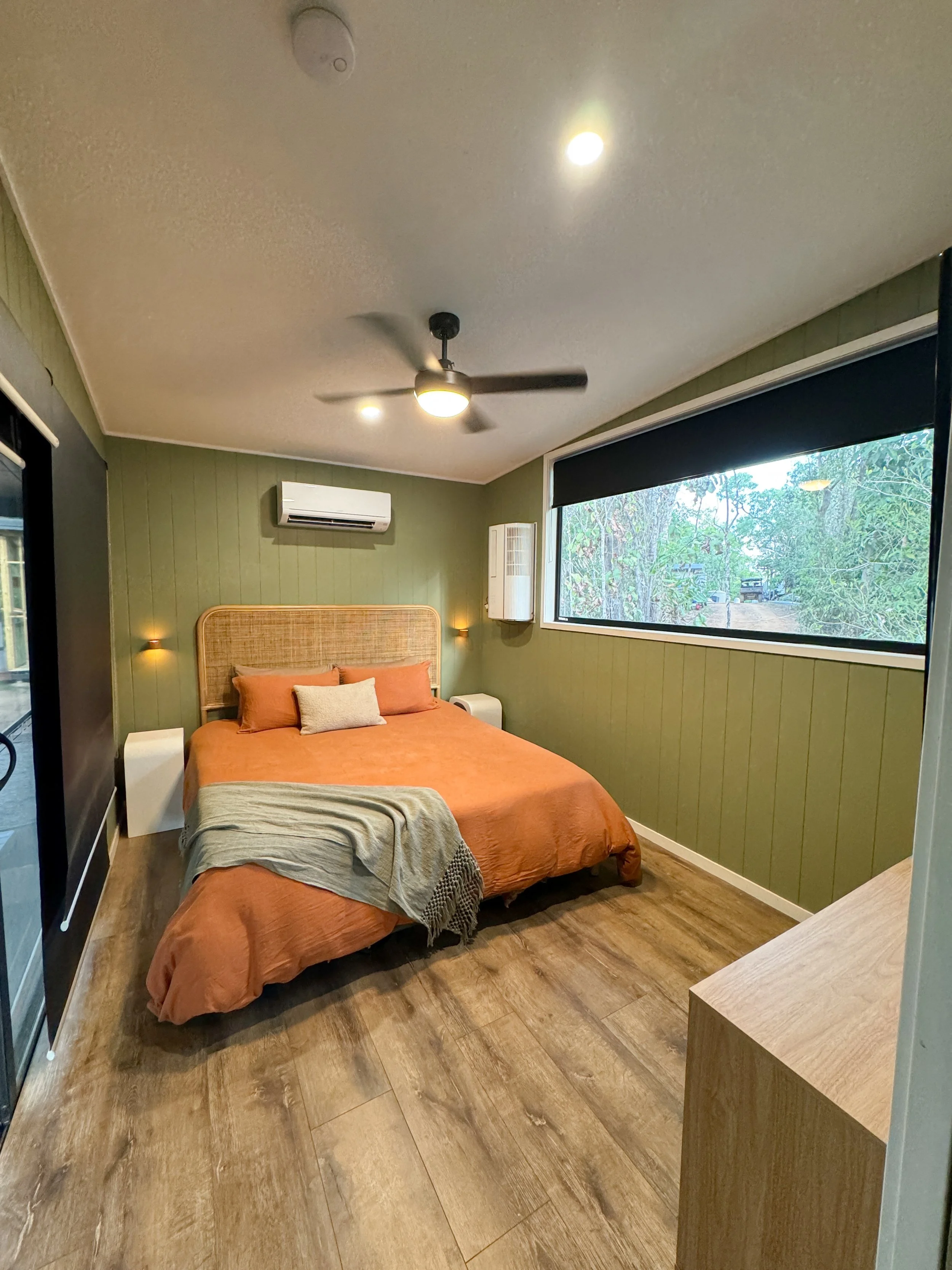 A bedroom with a green paneled wall, a large bed with an orange bedspread, matching pillows, and a small white pillow. There are wall-mounted small lamps on either side of the bed. A large window with a black roller shade shows an outdoor view of tre