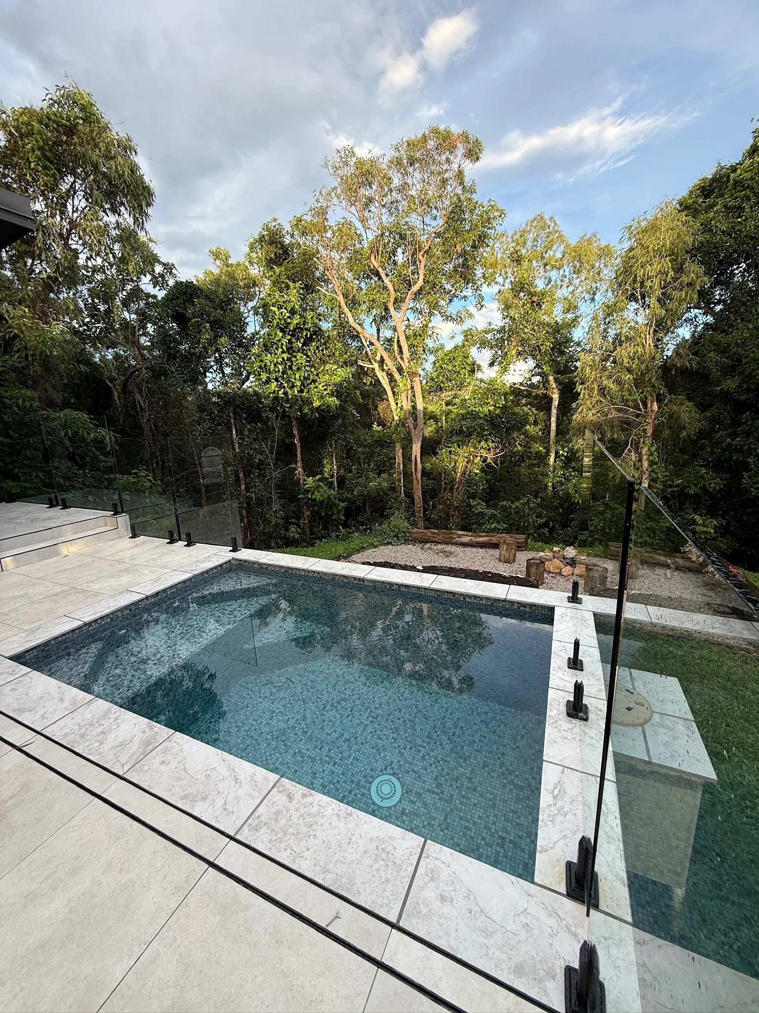 A modern rectangular swimming pool with clear water, surrounded by white stone tiles, adjacent to a lush green rainforest with tall trees, under a partly cloudy sky.