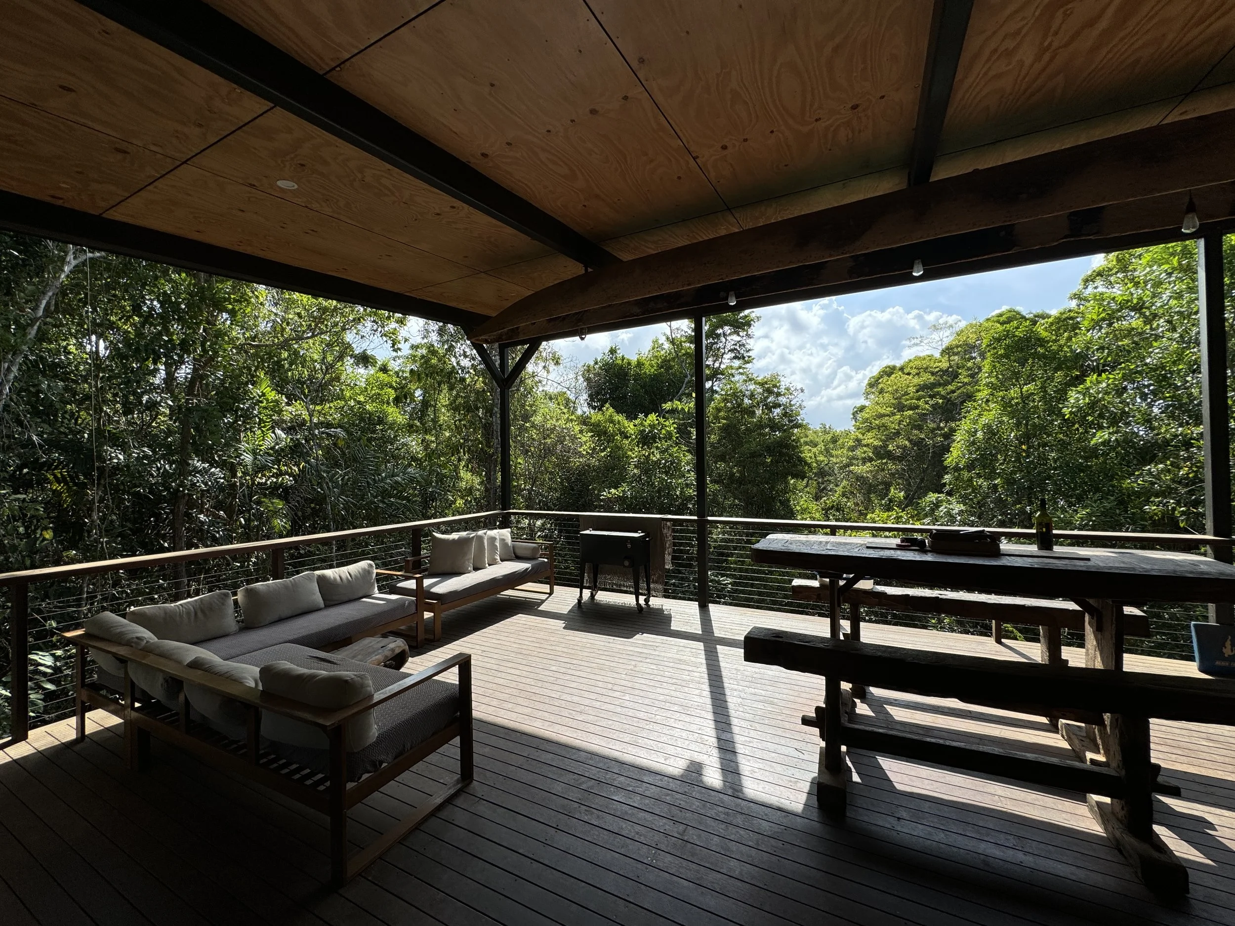 Covered outdoor deck with wooden flooring, outdoor furniture including sofas and a picnic table, and lush green trees in the background.