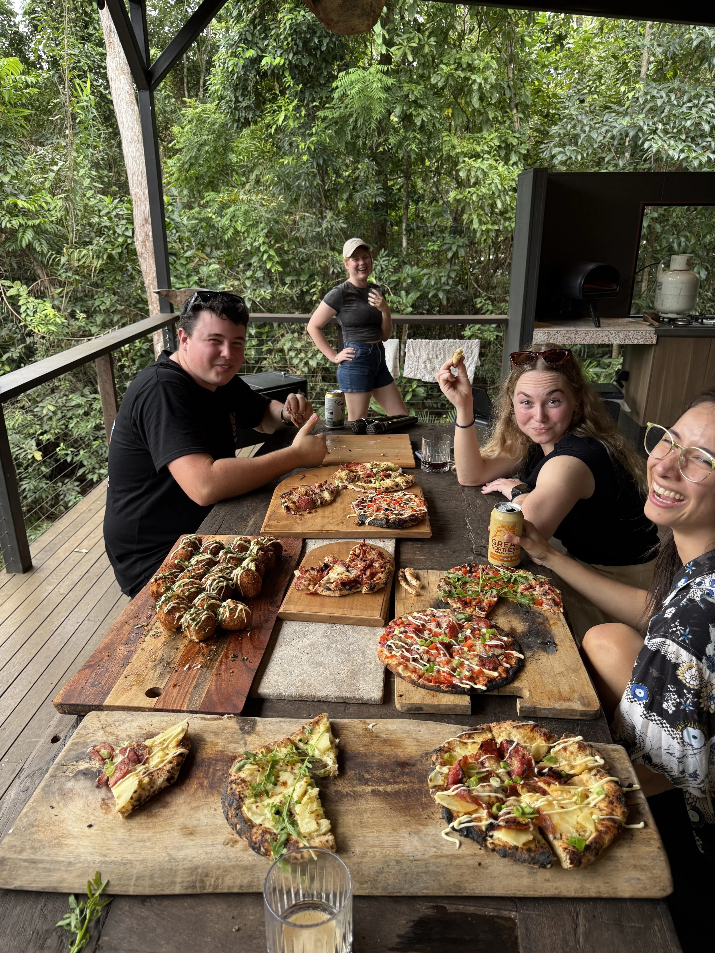 A group of five friends enjoying pizza and drinks at a wooden table on a covered deck surrounded by trees. They are smiling and laughing, with some holding pizza slices and drinks, and one person in the background standing near the railing.