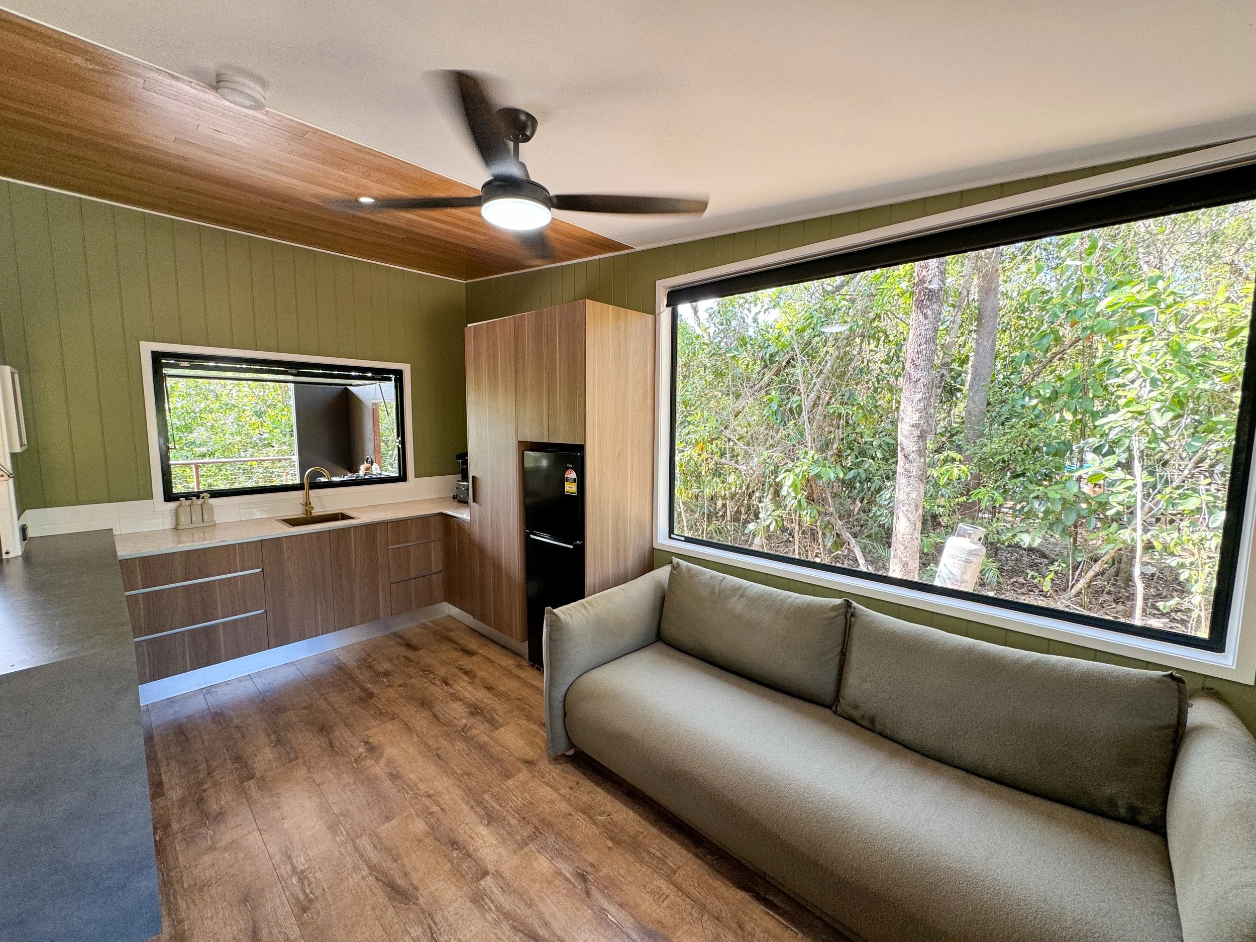Interior of a kitchen and living area with wood flooring, a green wall with vertical paneling, a black ceiling fan, a large window showing trees outside, a kitchenette with wood cabinets, a black refrigerator, and a beige sofa.