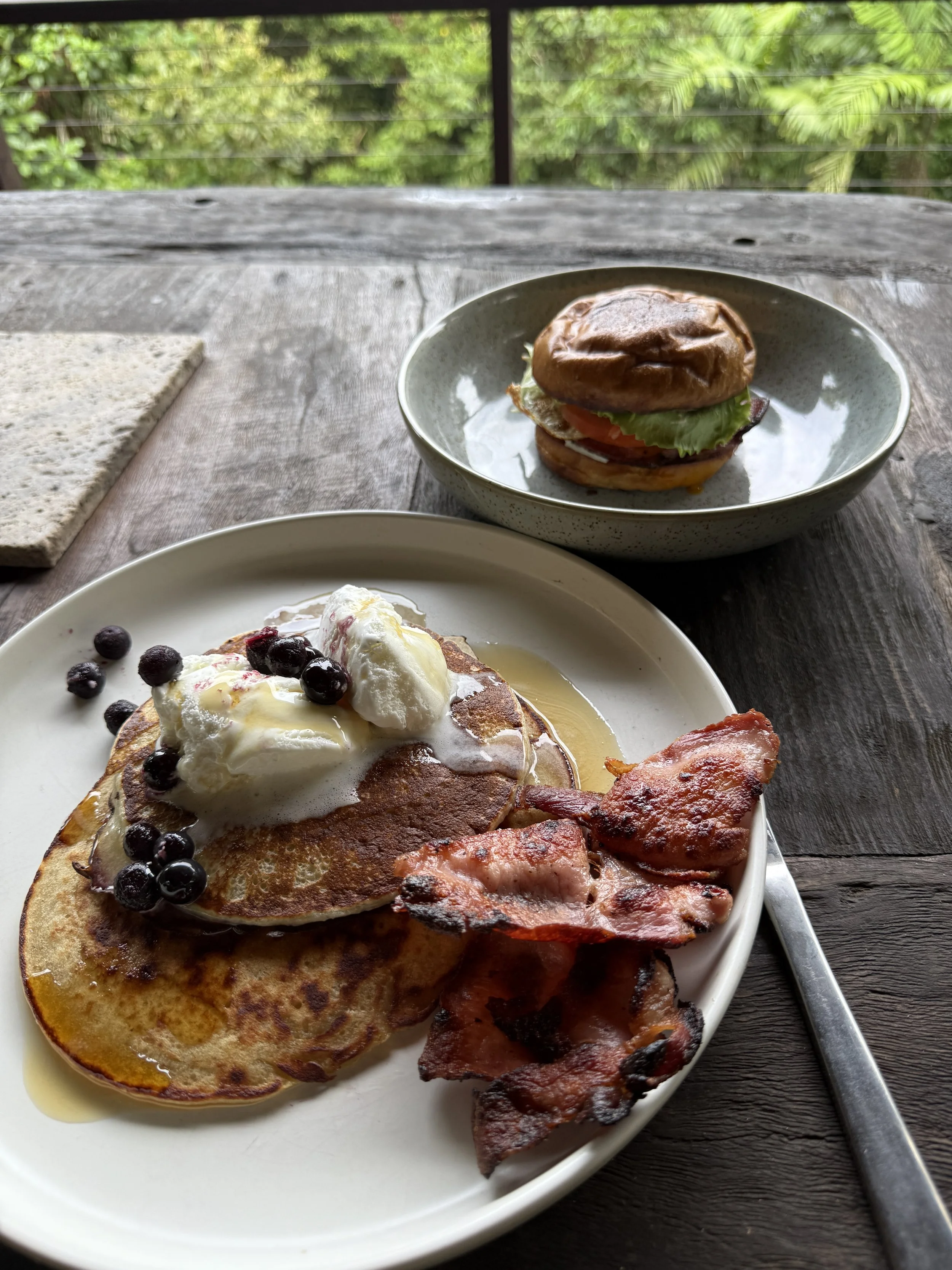 A plate of breakfast food with pancakes topped with whipped cream and blueberries, and crispy bacon strips on the side, in the foreground. In the background, a sandwich with lettuce, tomato, and possibly eggs in a bun, on a small plate.