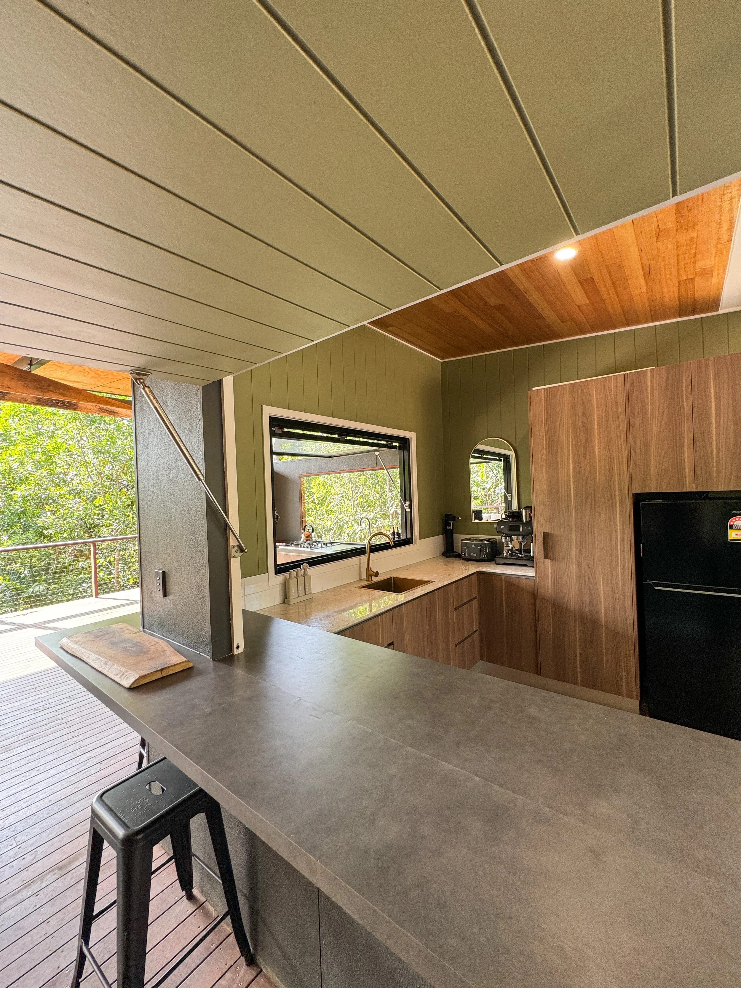 Modern kitchen with green and wood-paneled walls, a large window overlooking trees, a beige countertop, black refrigerator, and a breakfast bar with a black stool.