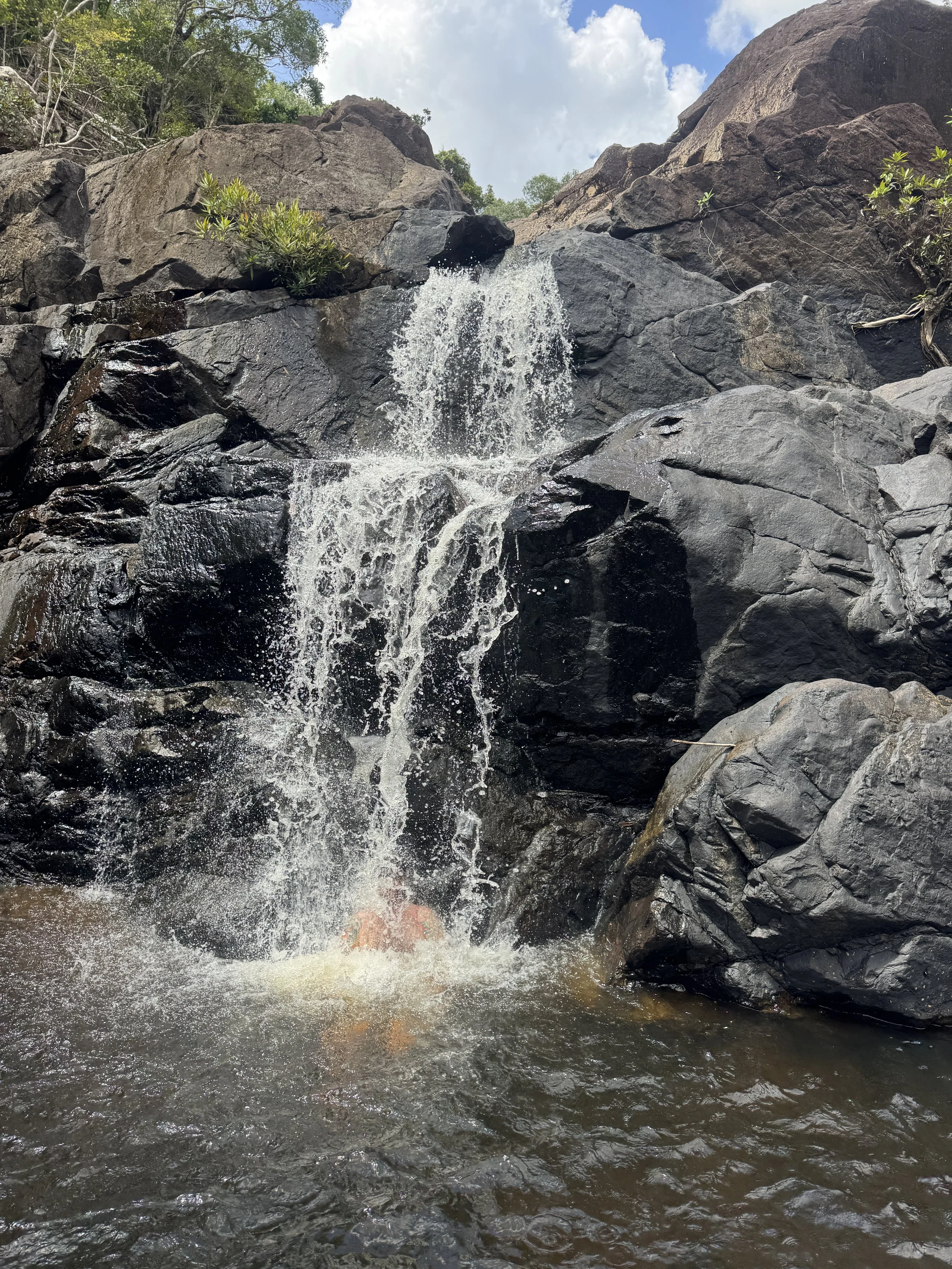 Small waterfall cascading over black rocks in a natural setting, with trees and a partly cloudy sky in the background.