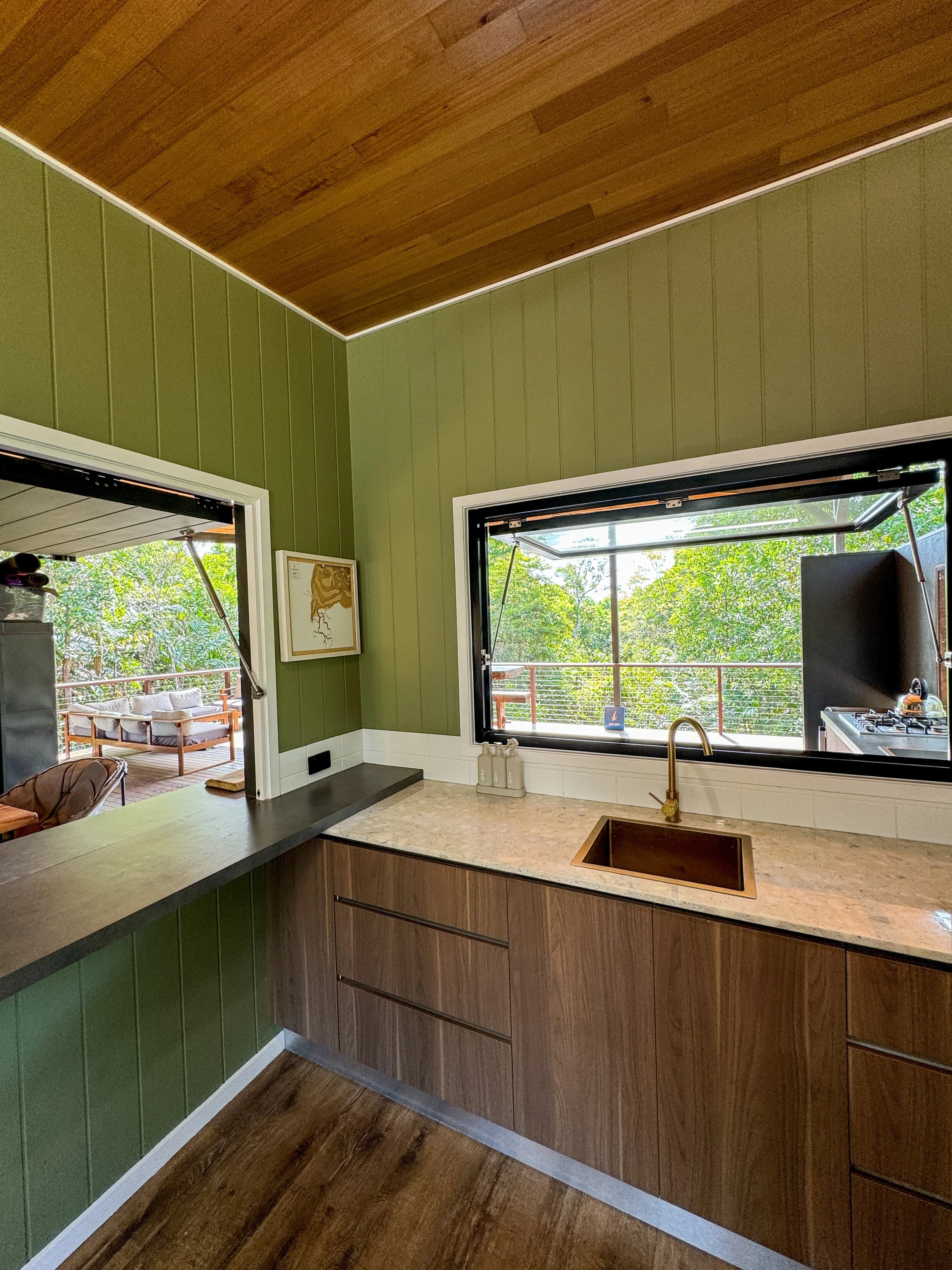 Modern kitchen with green paneled walls, wooden cabinets, marble countertops, a copper sink, and large open windows overlooking a deck with outdoor seating surrounded by trees.