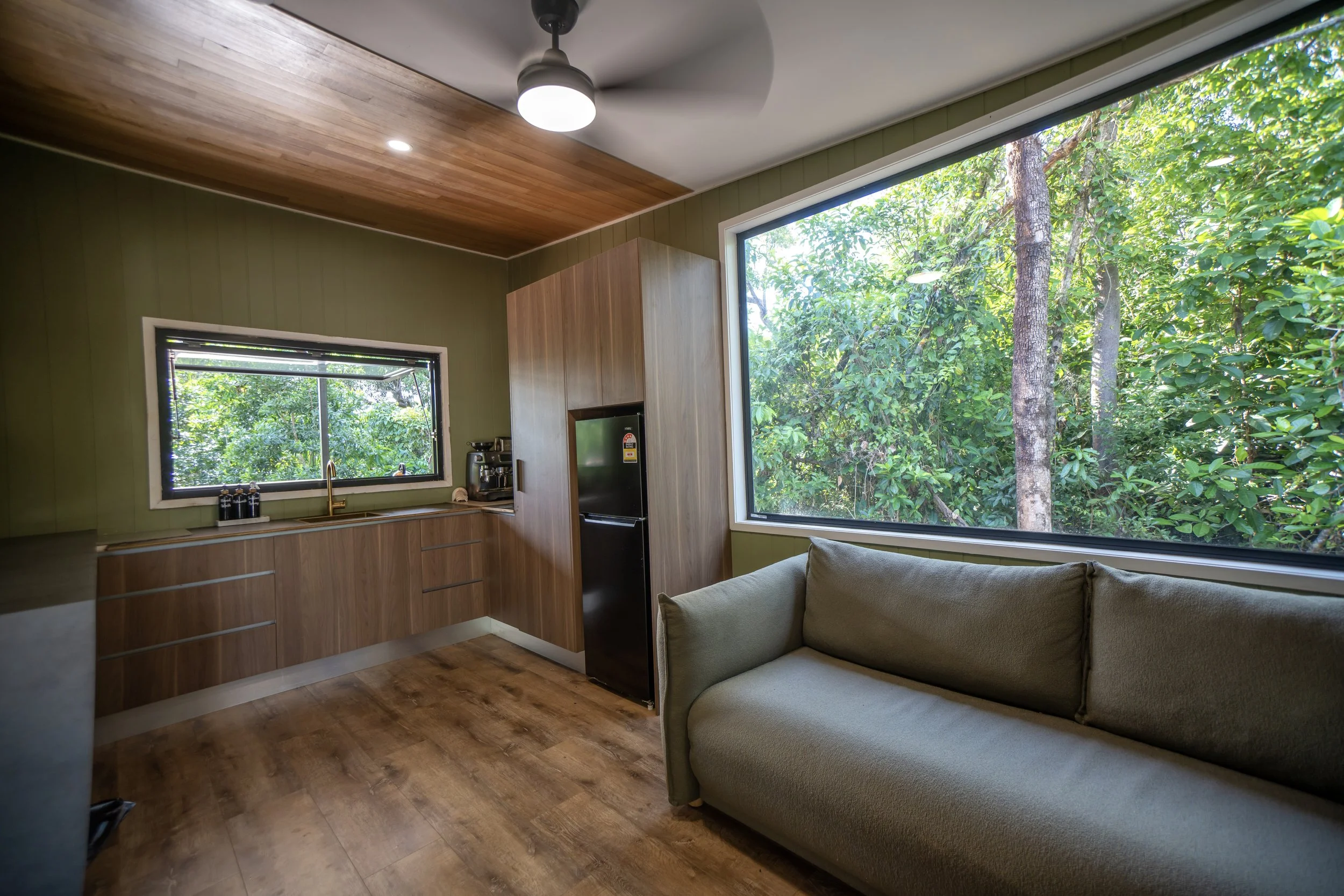 Interior of a cozy kitchen with wooden cabinets, a black refrigerator, a large window showing lush greenery, and a beige sofa beside the window.