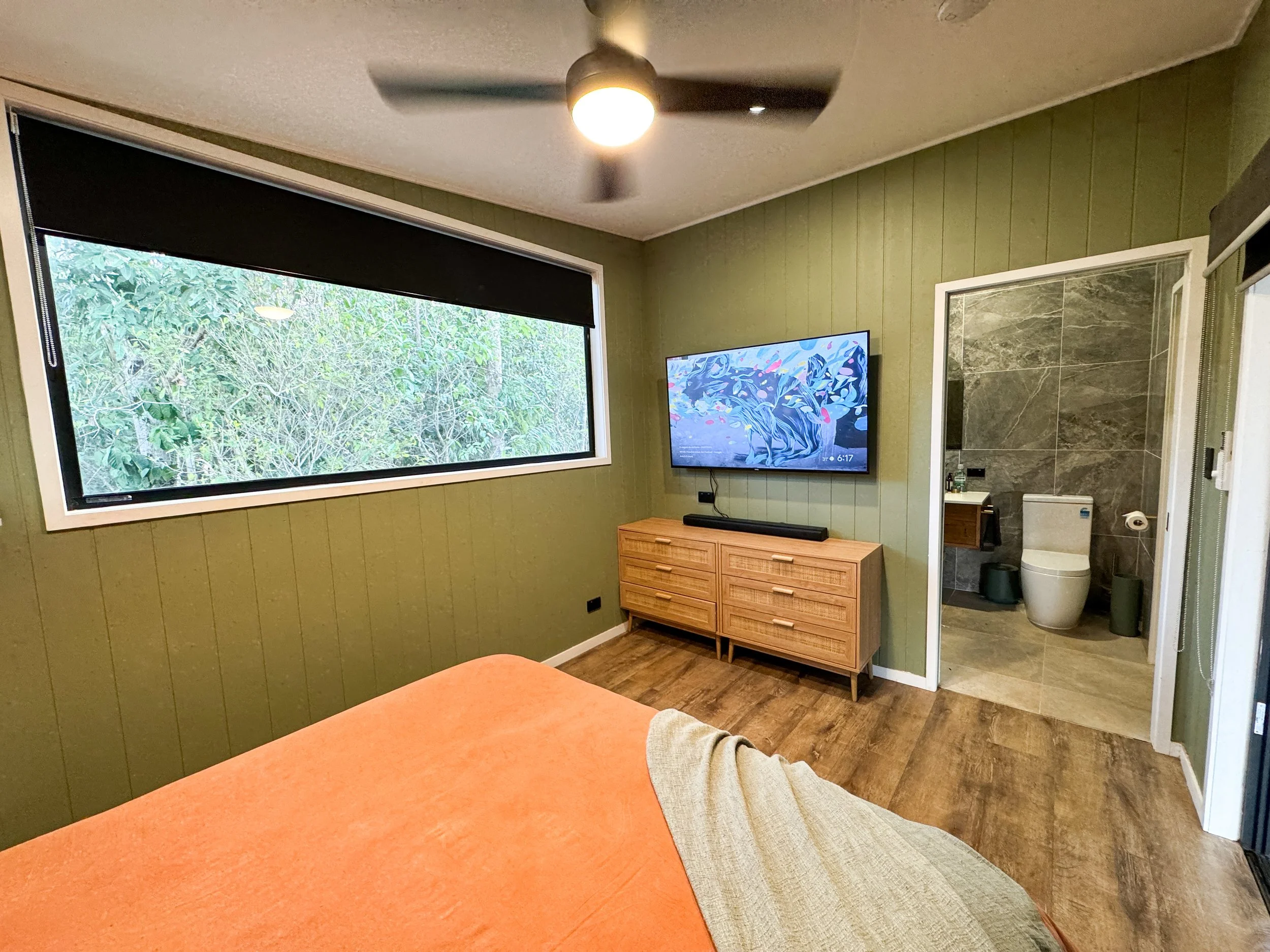 A cozy bedroom with a green paneled accent wall, a large horizontal window with a black shade, a flat-screen TV mounted on the wall, a wooden dresser beneath the TV, and a partially visible bed with an orange blanket and a beige throw. There is an en