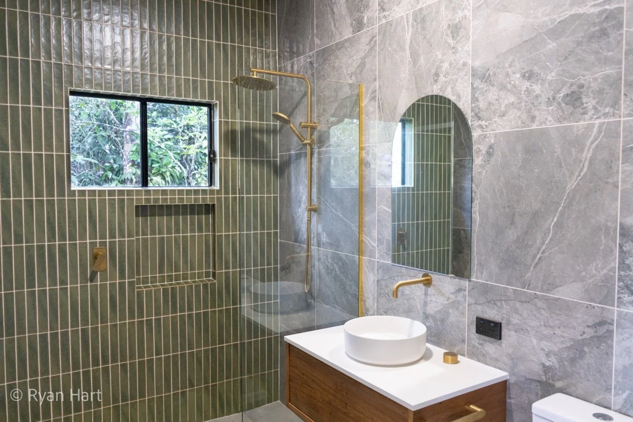 Modern bathroom with gray marble walls, green tile shower area, window with view of greenery, and a white vessel sink on a wooden vanity.