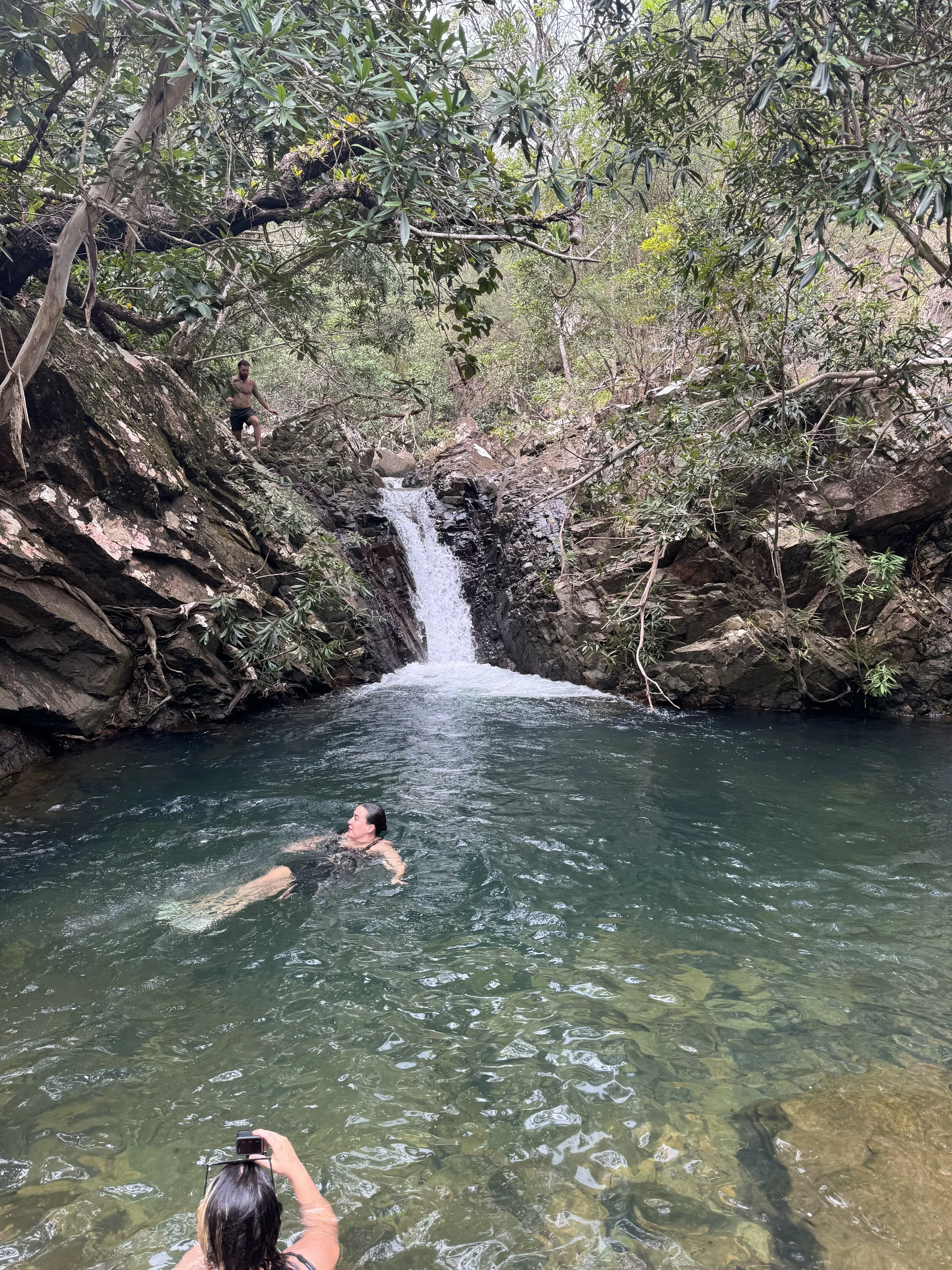 A woman floating in a small pool beneath a waterfall in a forested area, with a person taking her photo from the shore and a man standing on rocks near the waterfall.
