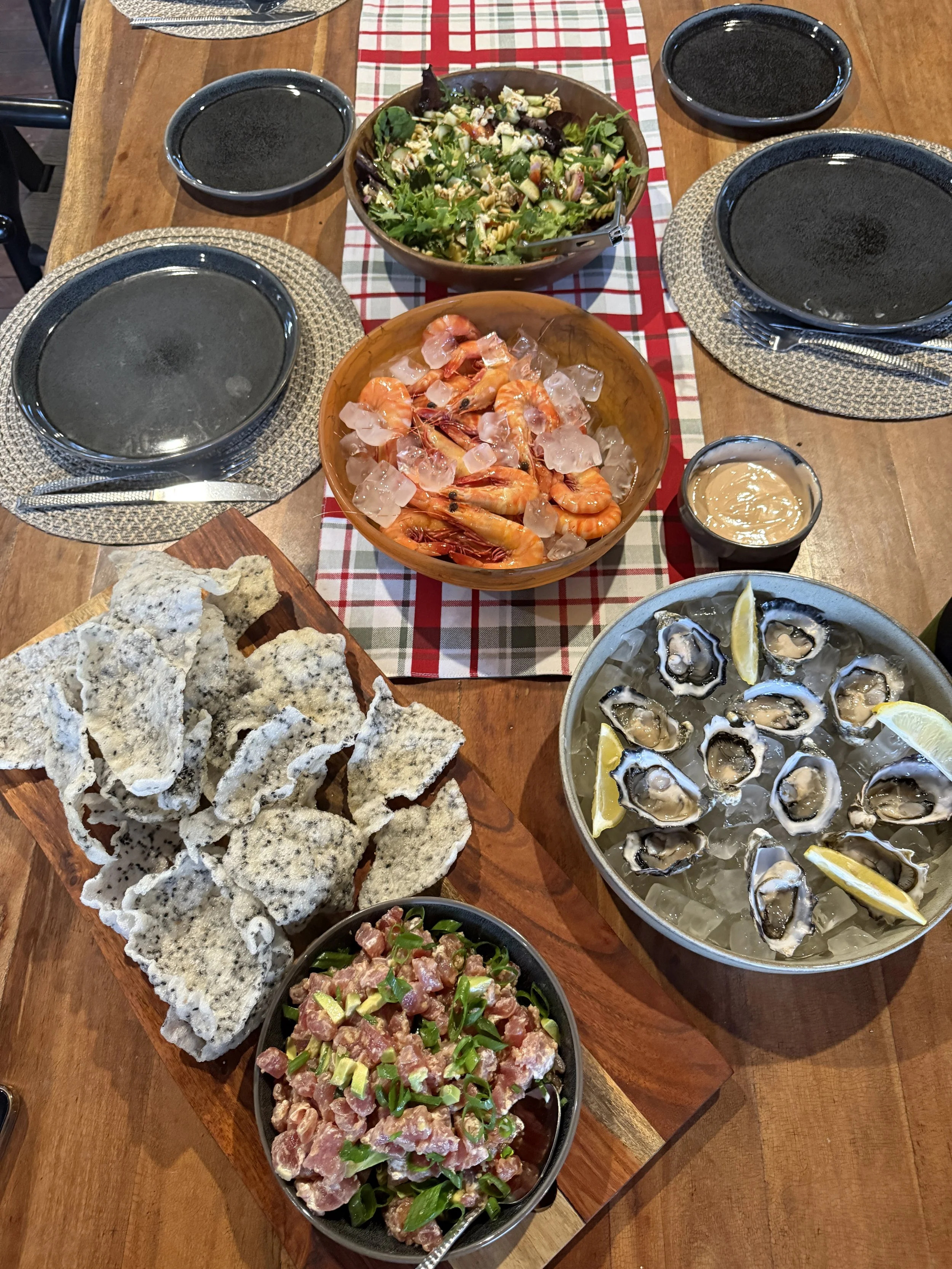 Seafood dinner spread with oysters on the half shell, shrimp on ice, a salad, beef tartare, crispy fried snacks, and dipping sauces on a wooden table.