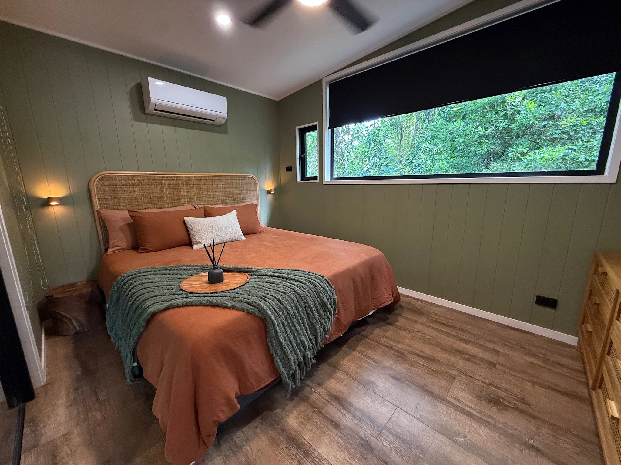 A bedroom with a green wooden wall, a large window showing greenery outside, a bed with rust coloured bedding, pillows, a green throw blanket, a wooden tray with a black diffuser, bedside lights, and an air conditioning unit on the wall.
