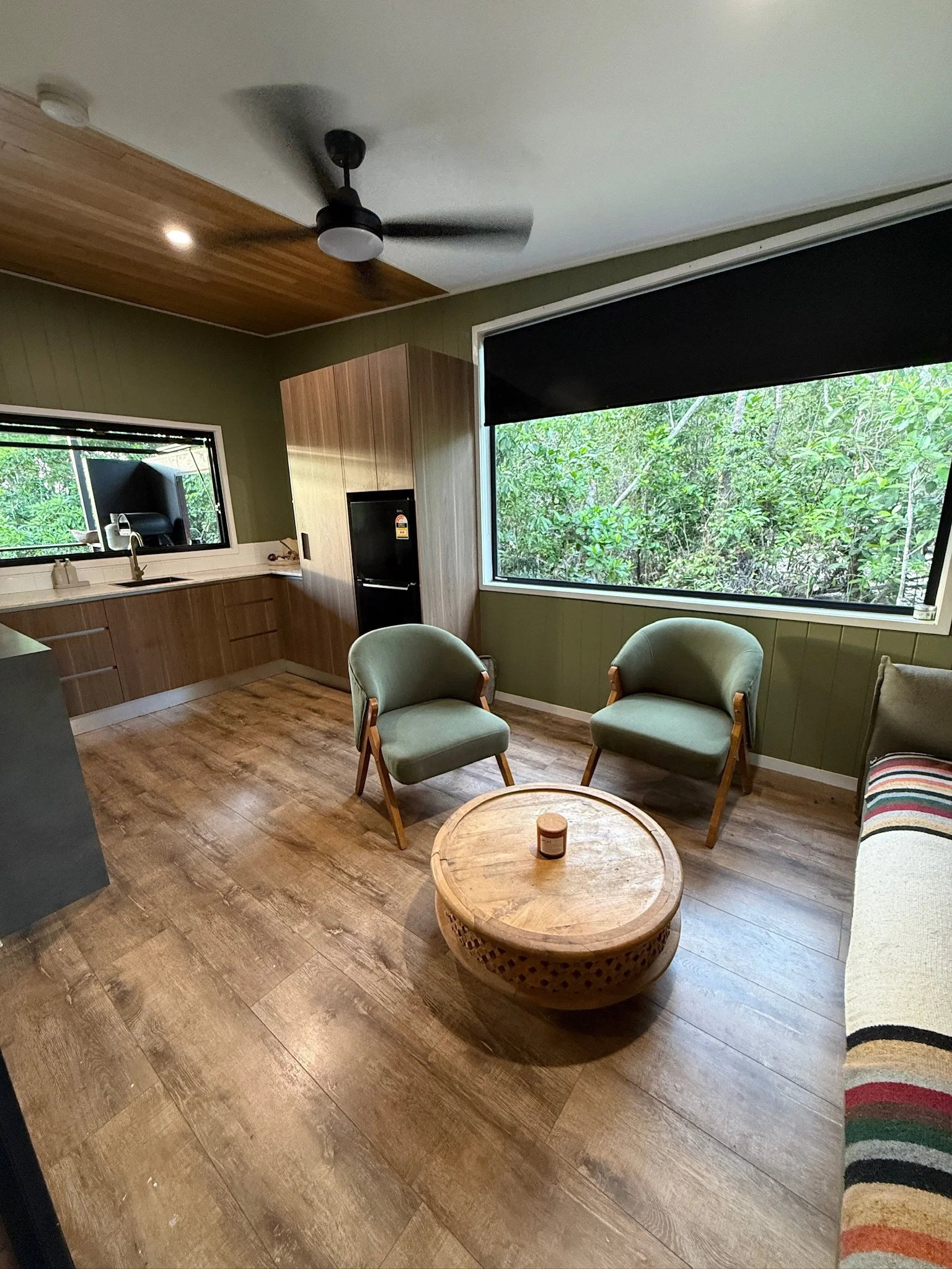 A cozy living room with two green upholstered chairs around a round wooden coffee table, a sofa with a colorful striped blanket, wooden flooring, large windows revealing green foliage outside, a ceiling fan, and a kitchen area with wooden cabinets an
