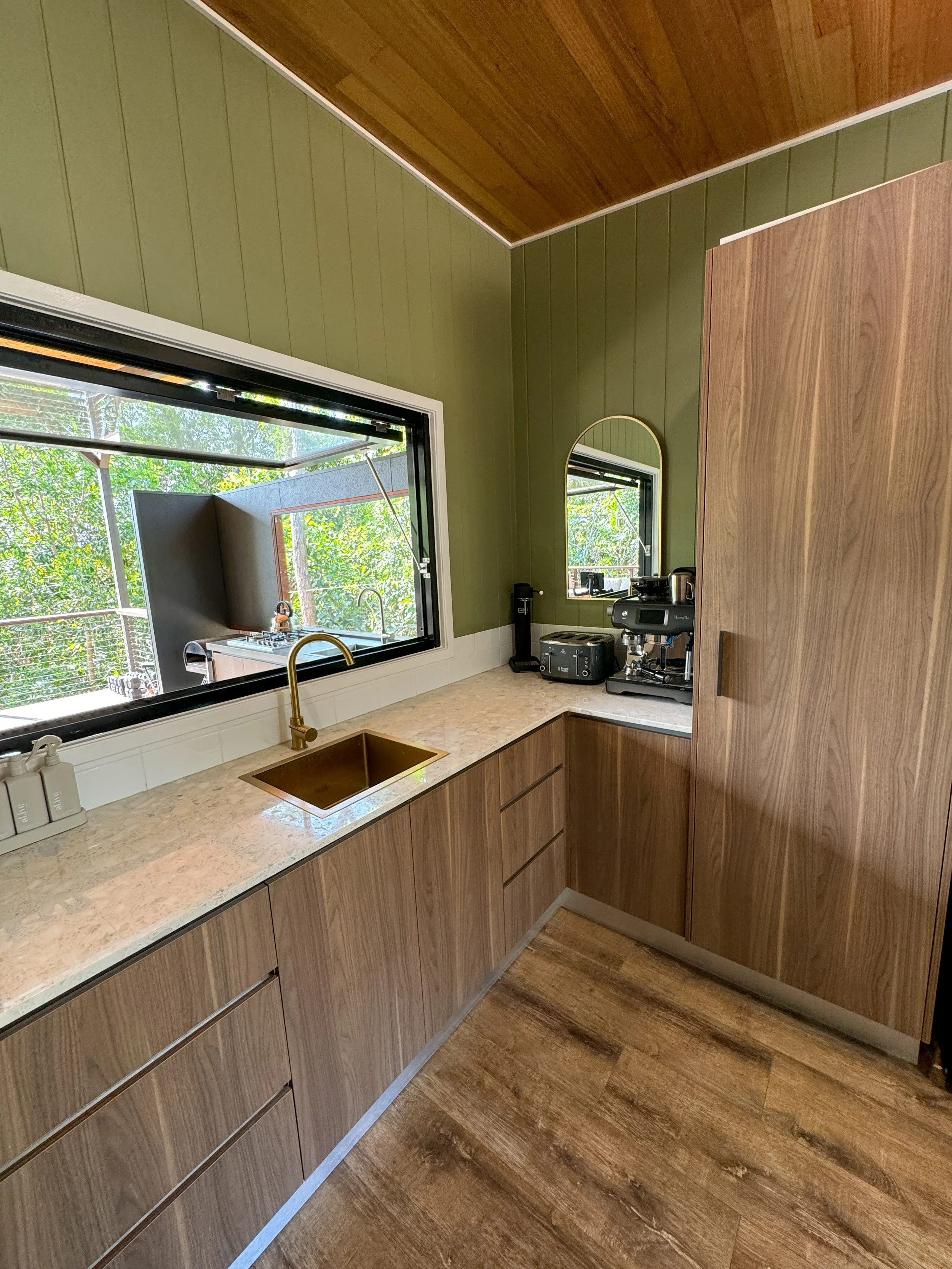 Kitchen with green and wooden walls, a sunny window, wooden cabinets, a gold sink, coffee machine, and a small mirror.