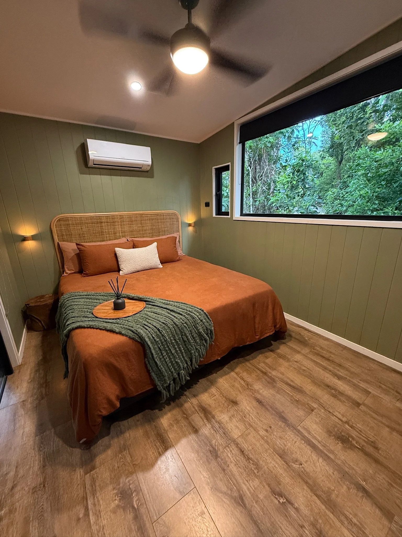 A bedroom with green paneled walls, a large window showing trees outside, a ceiling fan, a bed with an orange bedspread, brown pillows, a small white pillow, and a green knitted blanket. There are two wall-mounted lights above the bed and a small win