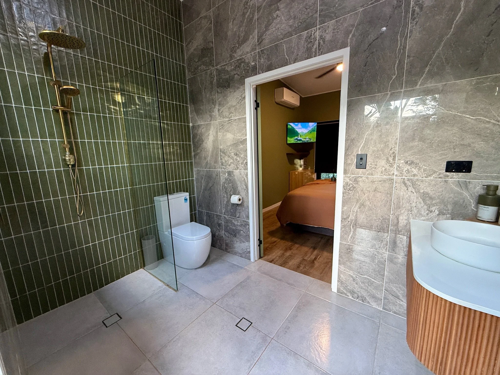 Modern bathroom with green tiled shower area, white toilet, wooden cabinet, and a bathroom sink with a round white basin, adjacent to a doorway leading to a bedroom with a bed, television, and air conditioning.