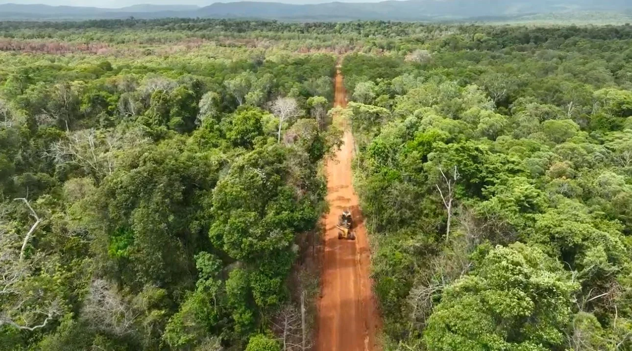Aerial view of a dirt road cutting through a dense green forest with construction vehicles on the road.
