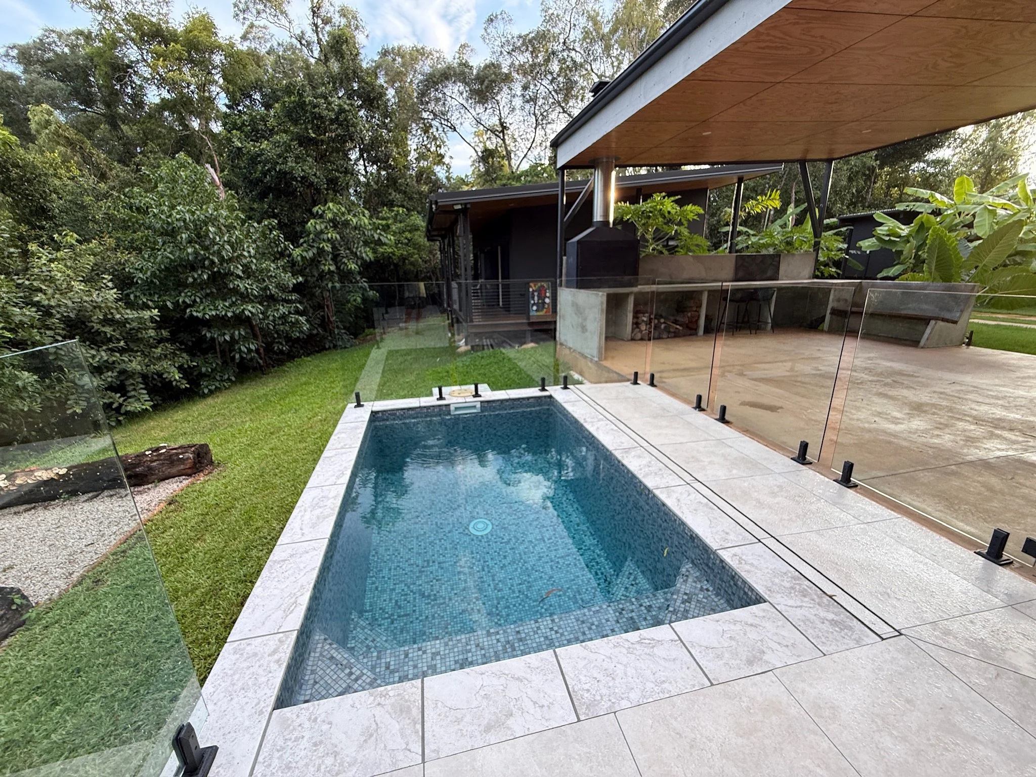 Small rectangular backyard swimming pool with a stone border, fenced with glass panels, next to a grassy area surrounded by the rainforest. A eco retreat with an outdoor patio and grilling area.