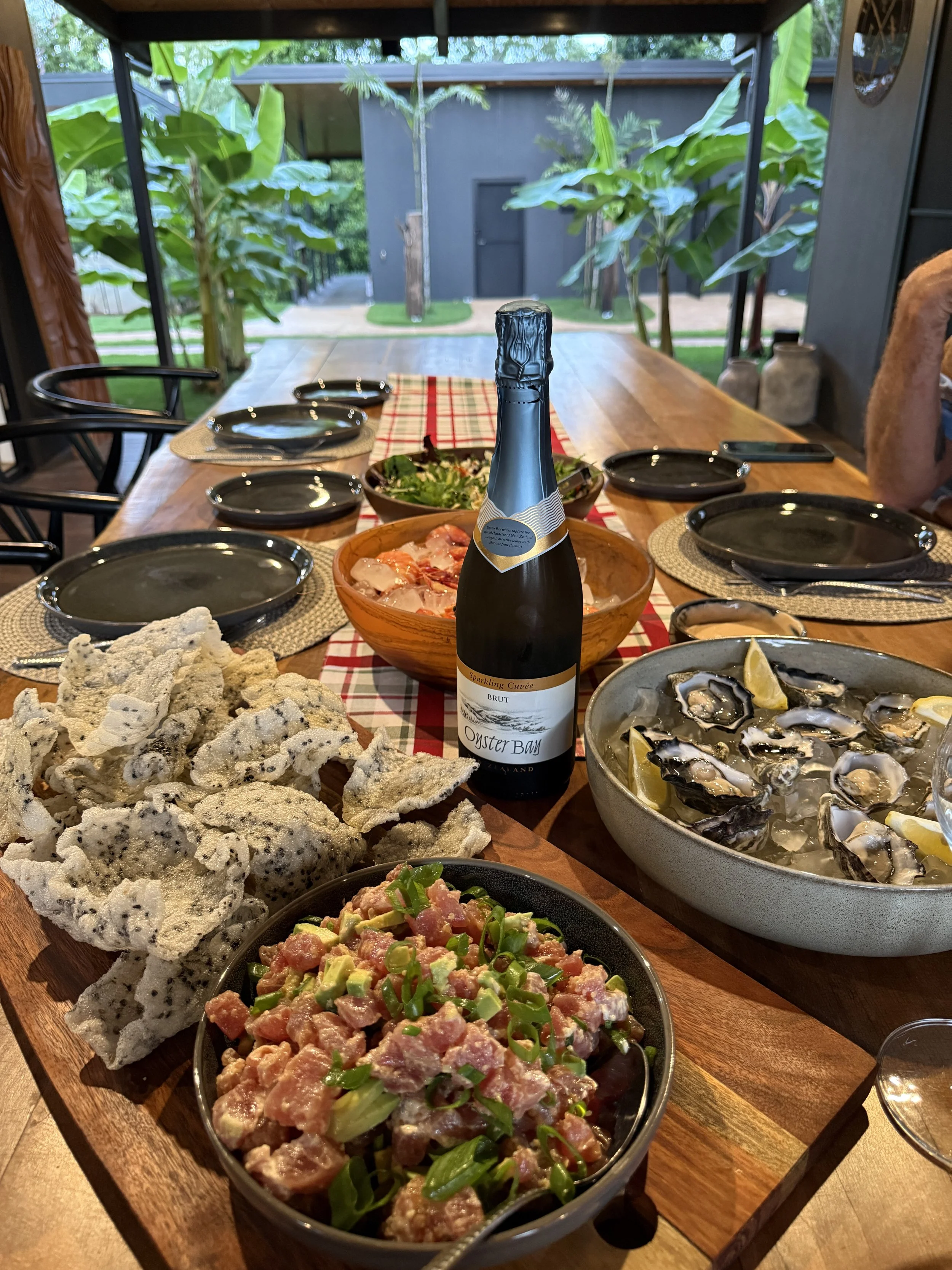 A wooden dining table set for a meal with plates, bowls of food including a bowl of oysters with lemon wedges, a bowl of chopped meat and green onions, a bowl of salad, and a pile of rice crackers with black sesame seeds. A bottle of sparkling wine i