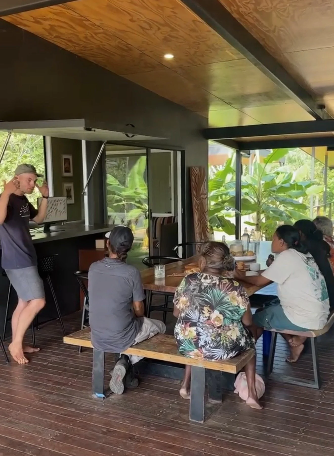 A group of people gathered around a wooden table inside a room with large windows and green foliage outside, with a man standing and speaking to them.