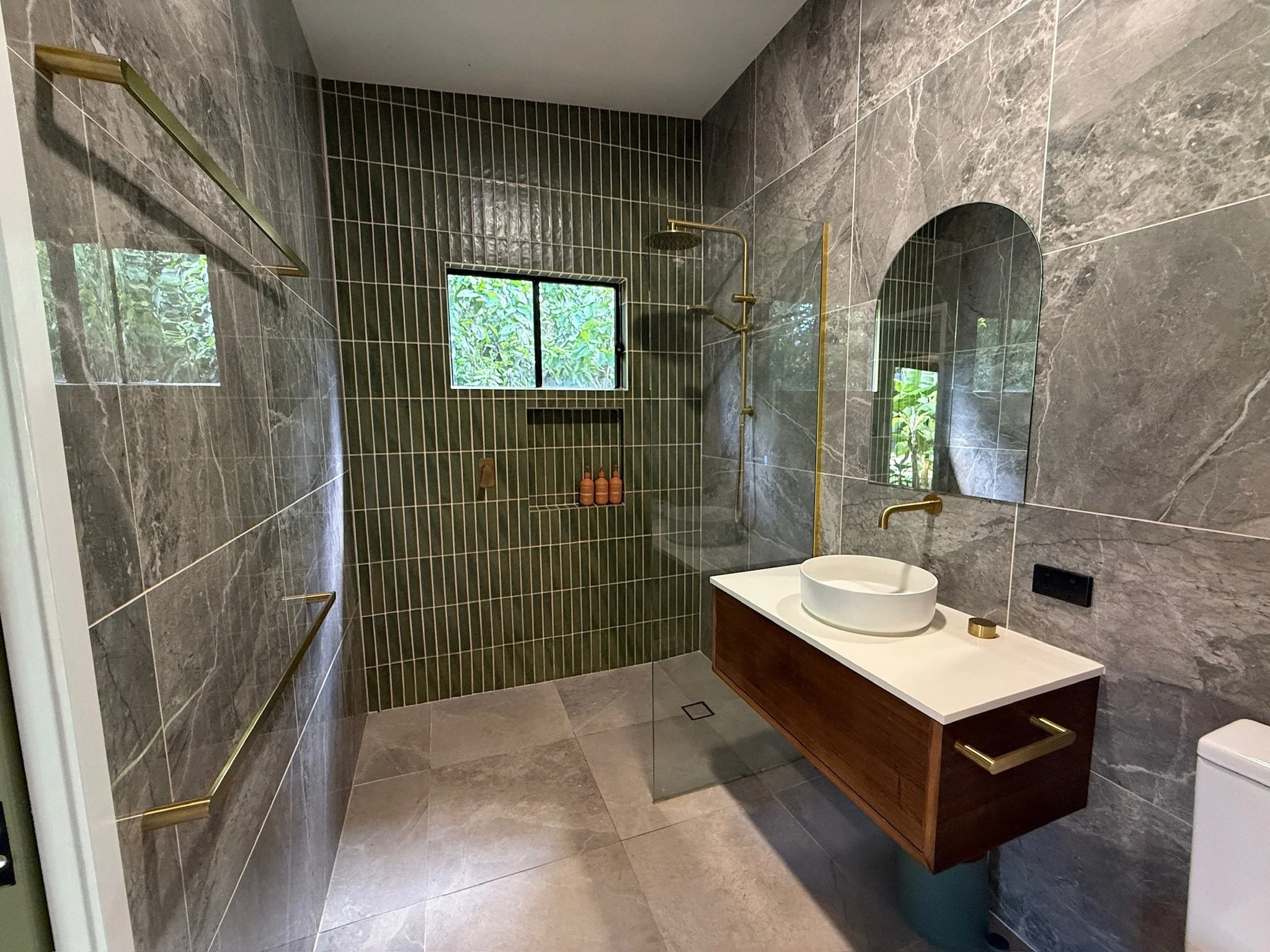 Modern bathroom with gray marble walls, a green tiled shower area with two small windows, gold fixtures, a wooden vanity with a vessel sink, and a mirror.